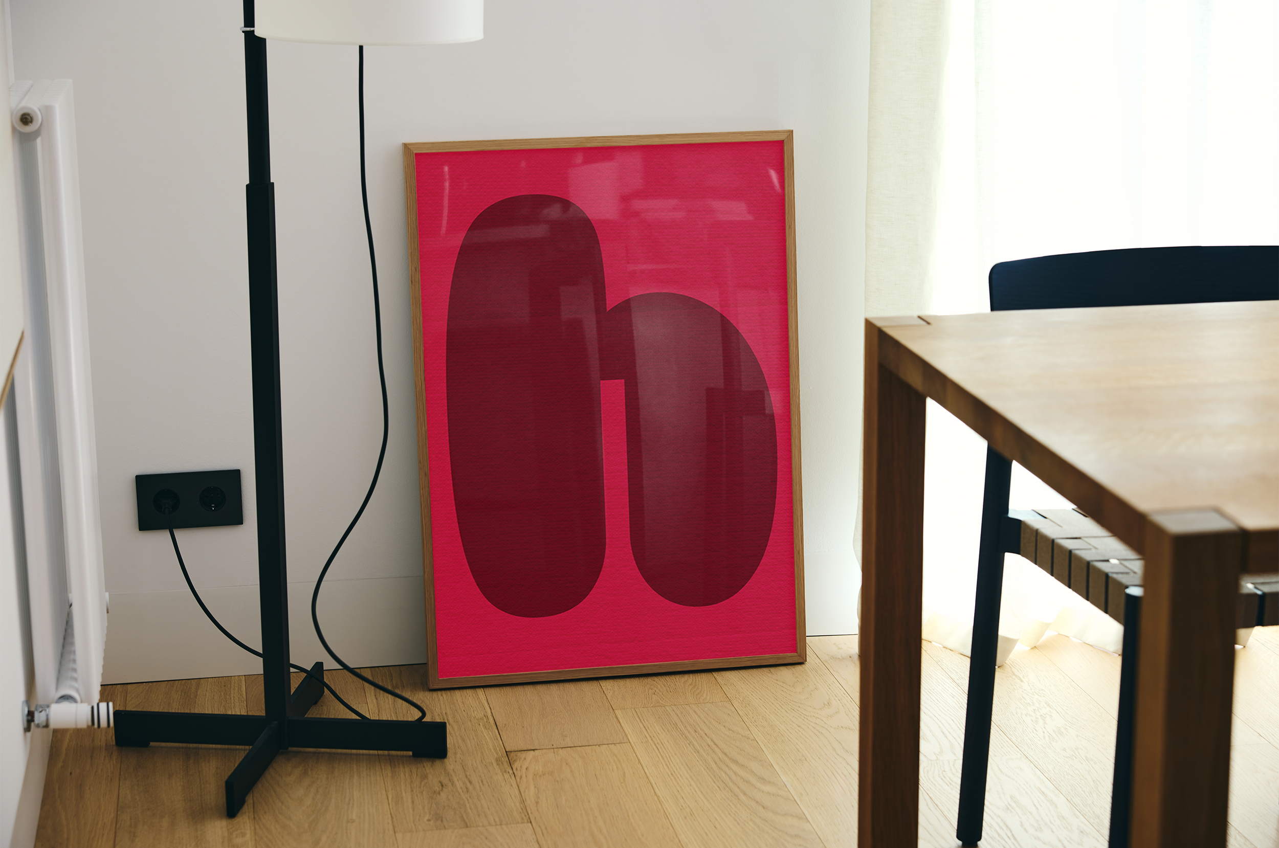 A light wood-framed abstract print with two large, textured maroon oval shapes on a bright pink background, resting on a light wooden floor next to a black floor lamp and a wooden table.
