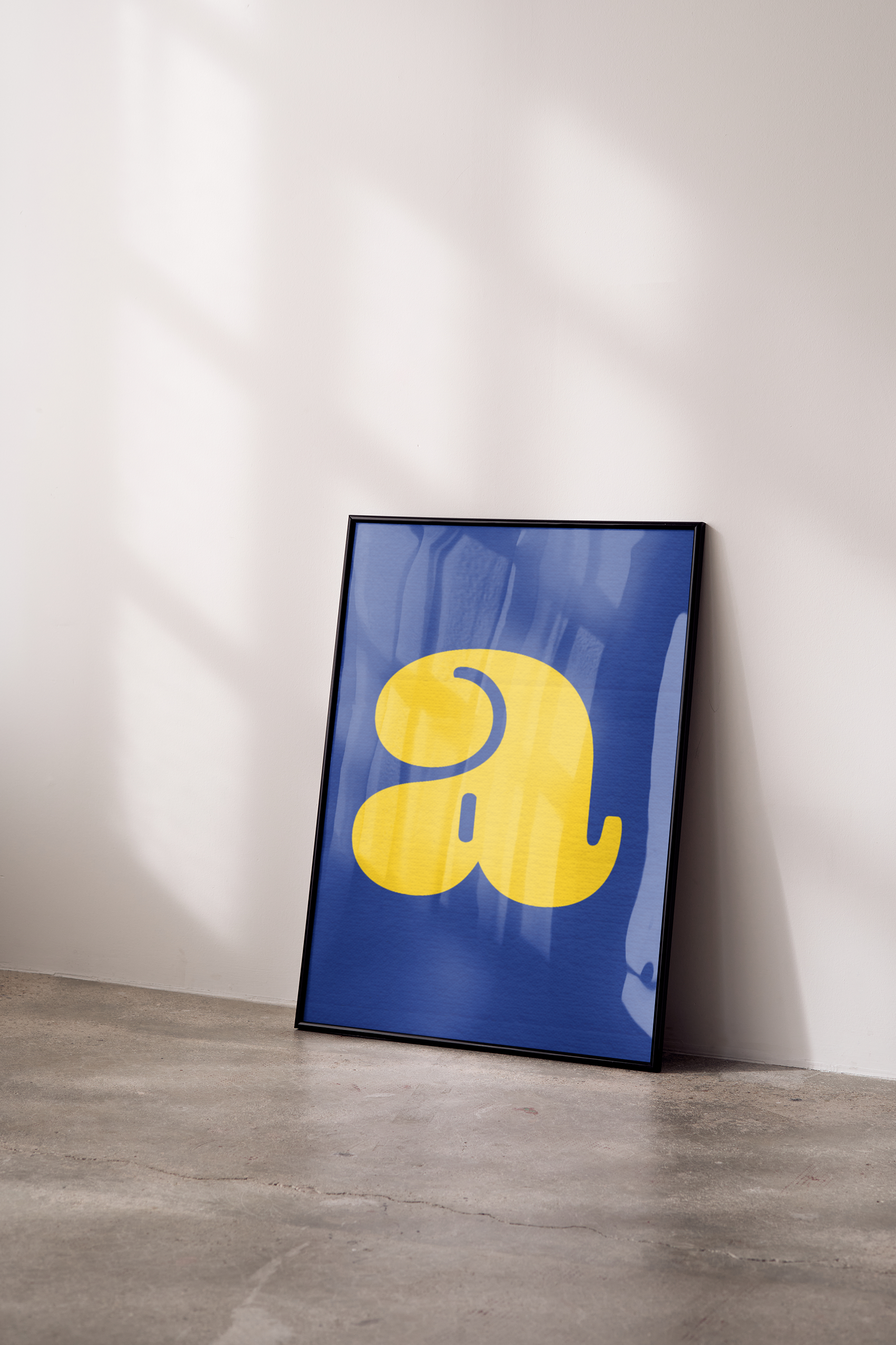 A framed poster with a large, fluid yellow lowercase letter 'a' on a blue textured background, leaning against a white wall in a sunlit space.