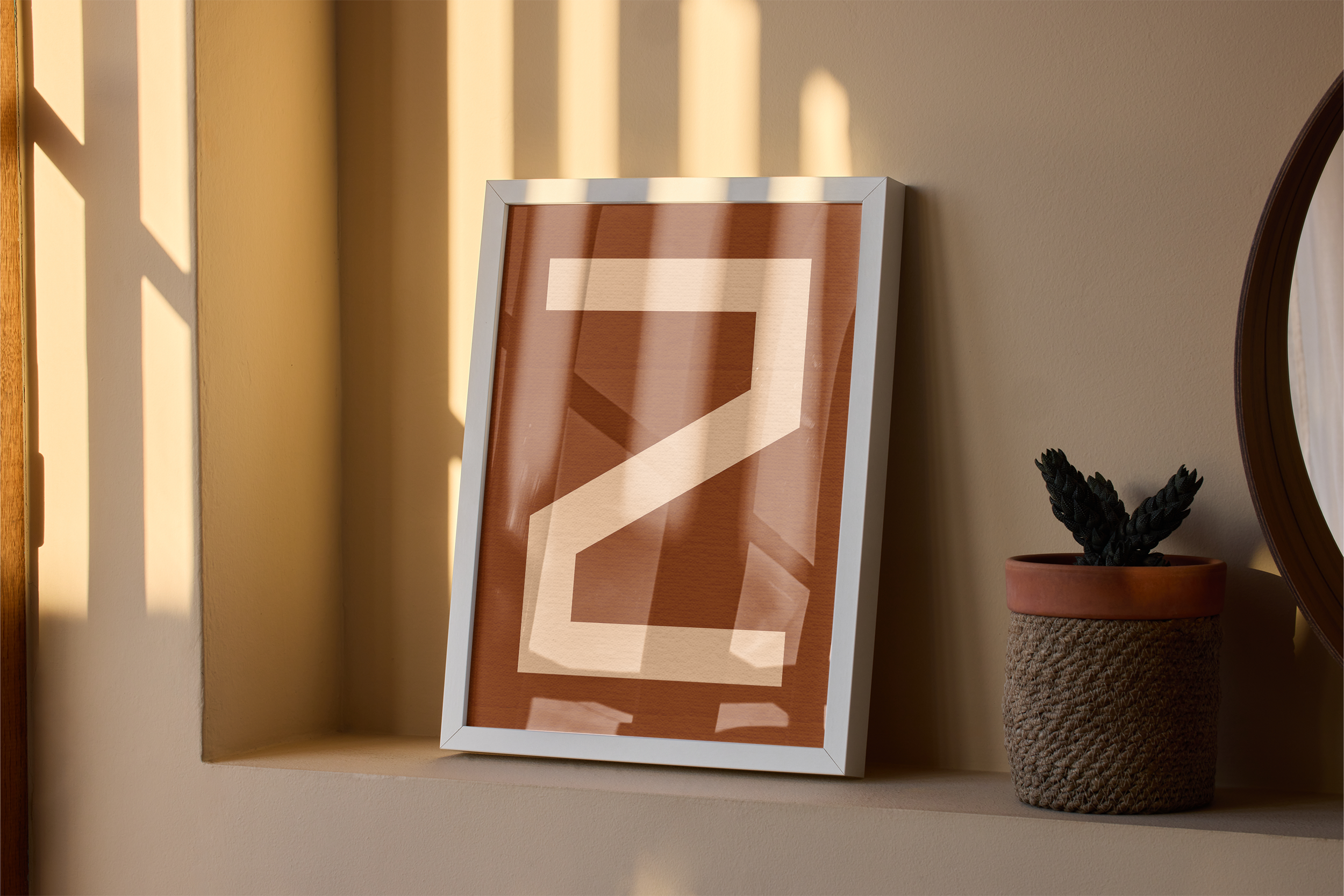 A white framed print featuring a large, abstract letter 'Z' in white on a brown background, resting on a beige shelf with a small potted plant and a round mirror nearby, bathed in sunlight and shadow stripes.