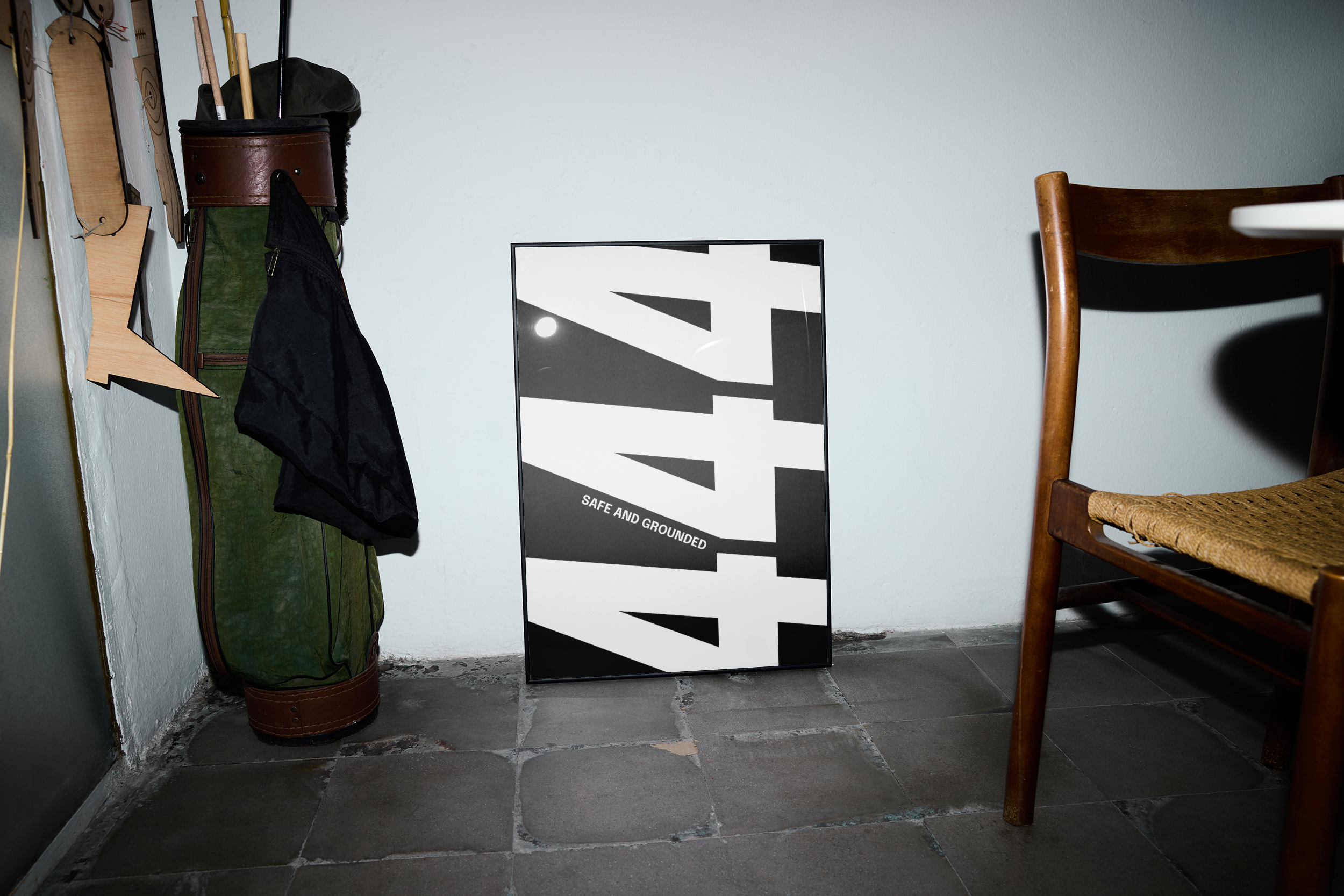 Black framed poster with the repeating number '444' in a black and white design with the text "SAFE AND GROUNDED," leaning against a light wall on a tiled floor next to a vintage golf bag and a wooden chair.