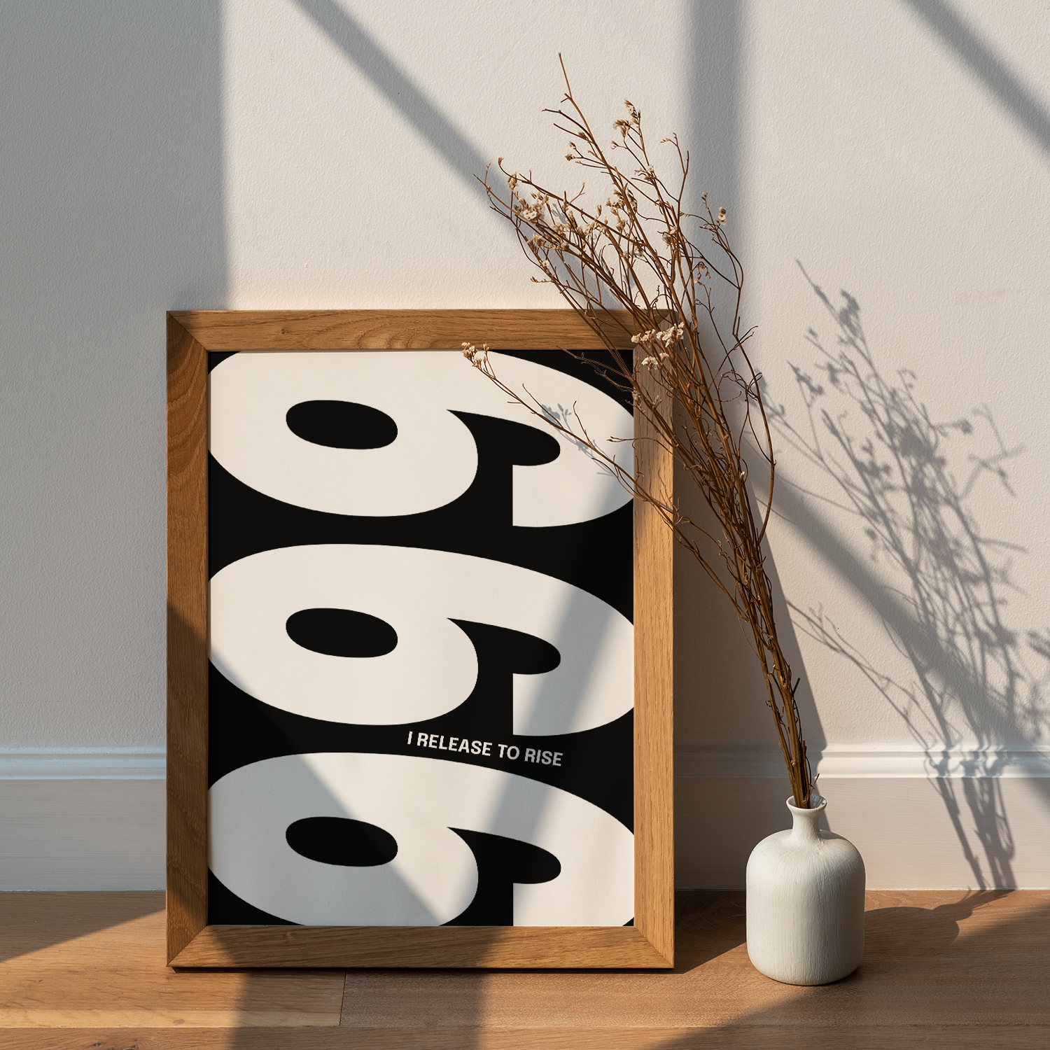 Wooden framed poster with the repeating number '999' in a black and white design with the text "I RELEASE TO RISE," leaning against a white wall on a wooden floor next to a white vase with dried branches.