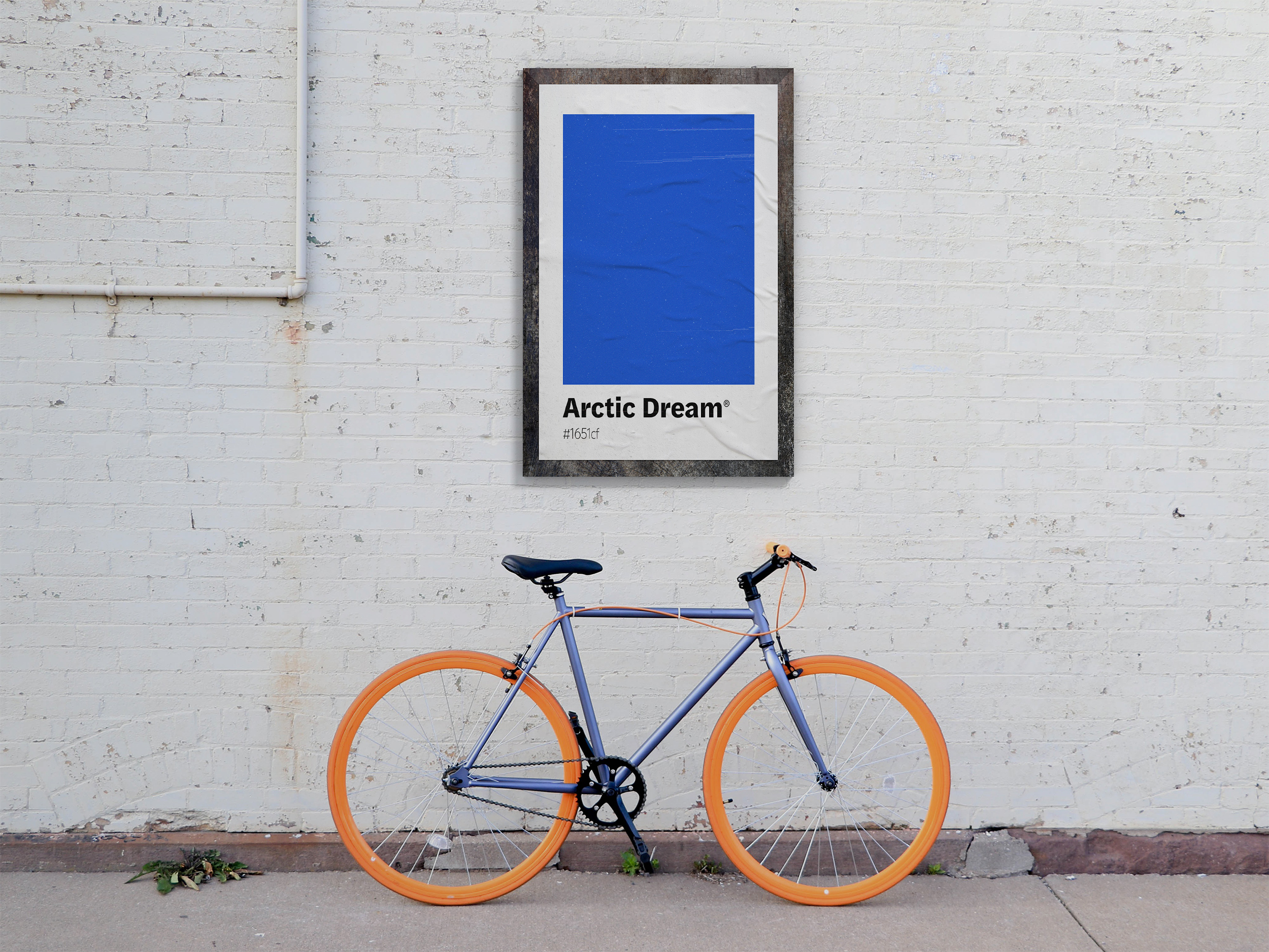 Framed print featuring a large block of bright blue labeled "Arctic Dream," hanging on a white brick wall above a gray bicycle with bright orange tires.