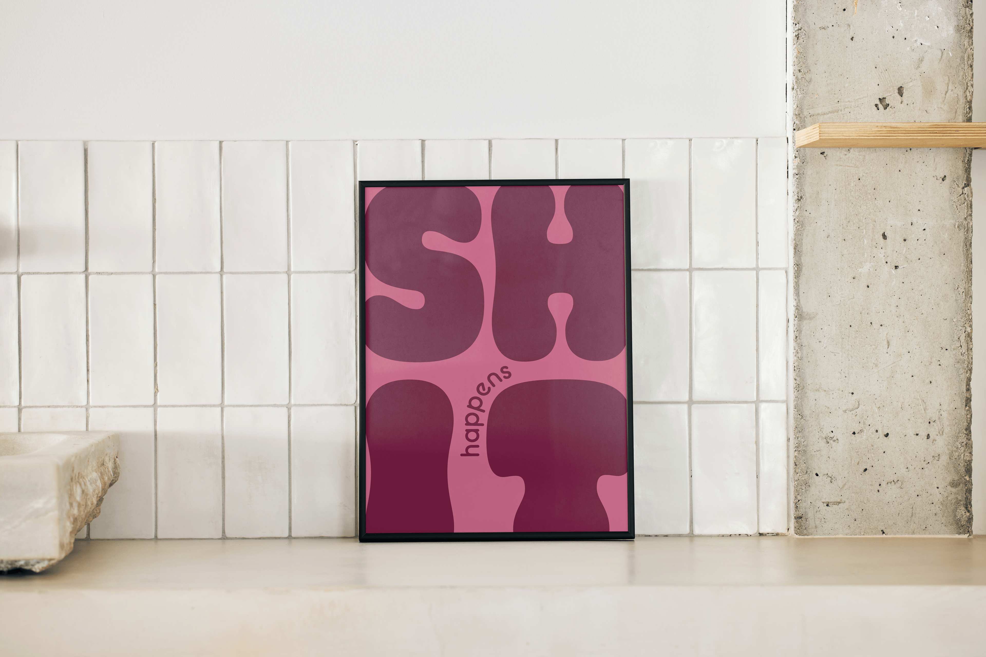 A framed art print with the text "SHIT happens" in large, rounded, dark pink letters on a lighter pink background is propped on a white tiled surface. The print is centered in the frame. To the left, a corner of a light-colored, rough-hewn sink is visible. To the right, a raw concrete pillar has a light wooden shelf extending from it. The background features white rectangular tiles.