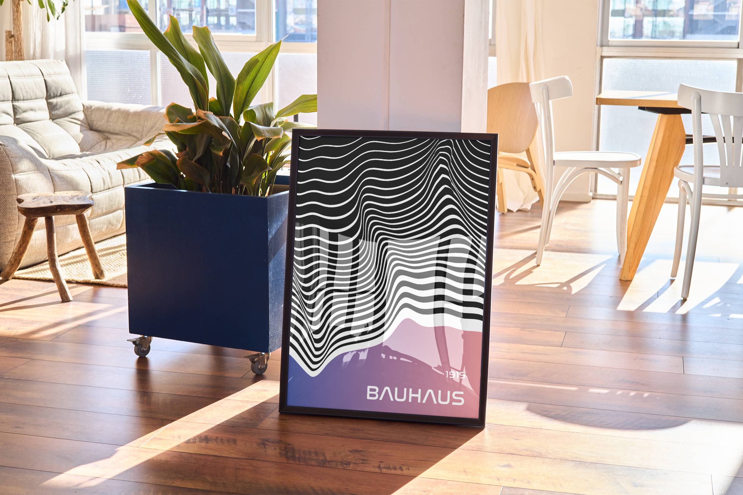 A black-framed modern art poster with abstract wavy lines and "BAUHAUS" text, resting on a wooden floor in a bright living room next to a blue planter.