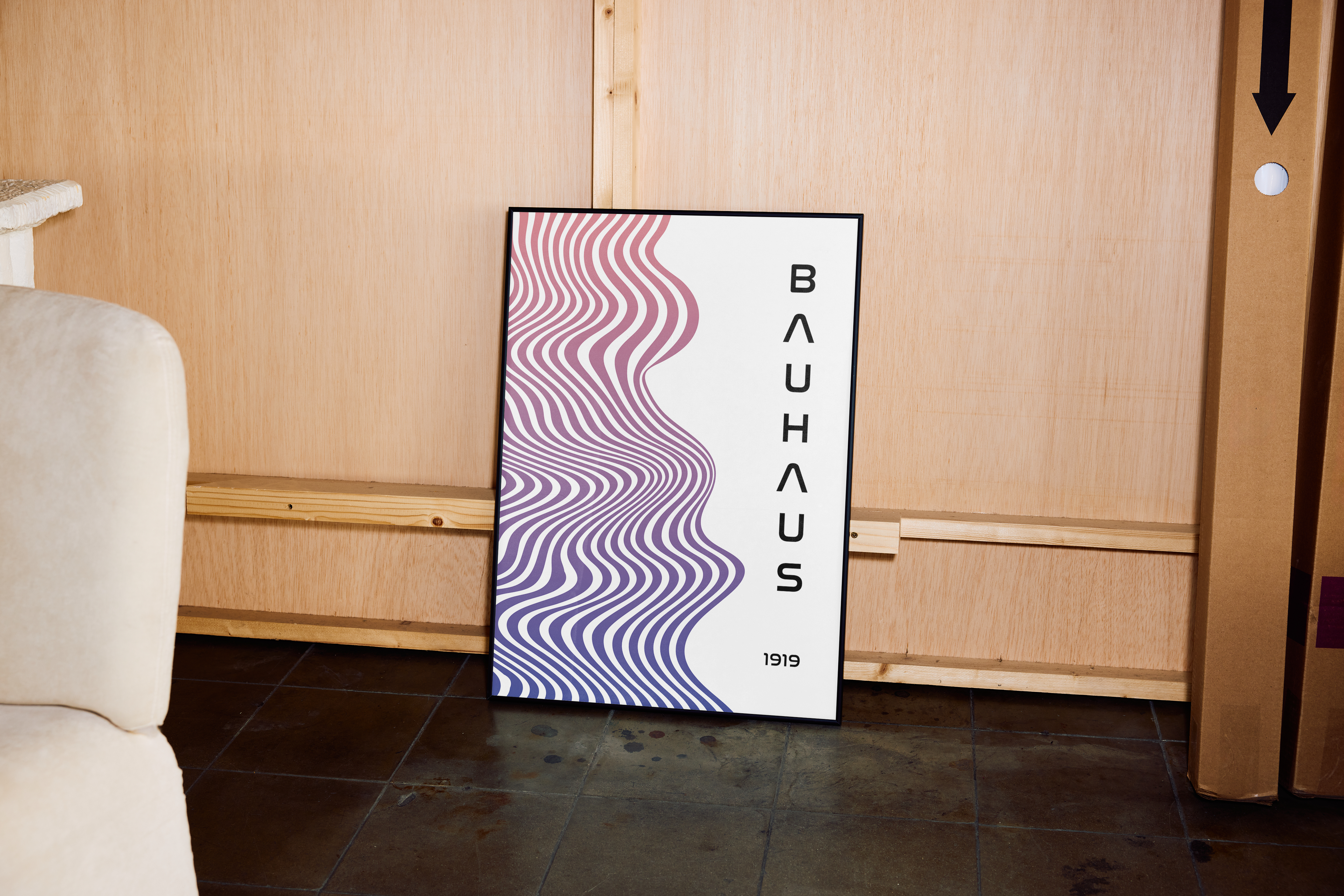A framed modern art poster with the word "BAUHAUS" vertically on the right and an abstract design of undulating, wavy lines in purple and pink gradients on the left, leaning against wooden wall paneling in a corner next to a beige chair and cardboard boxes.