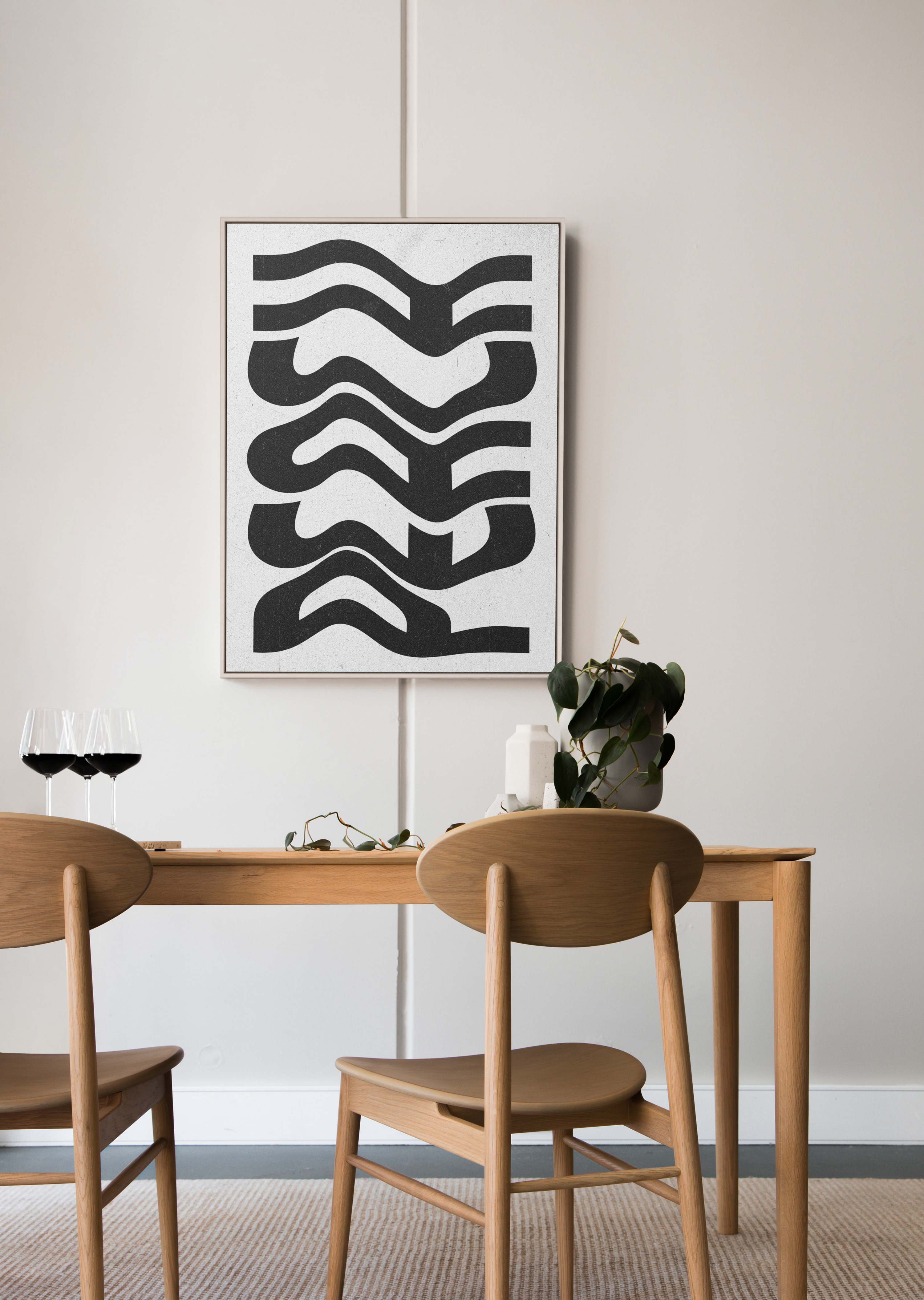 A gold-framed abstract art poster with thick, wavy black lines on a white textured background, hanging above a simple wooden dining table with two chairs and wine glasses.