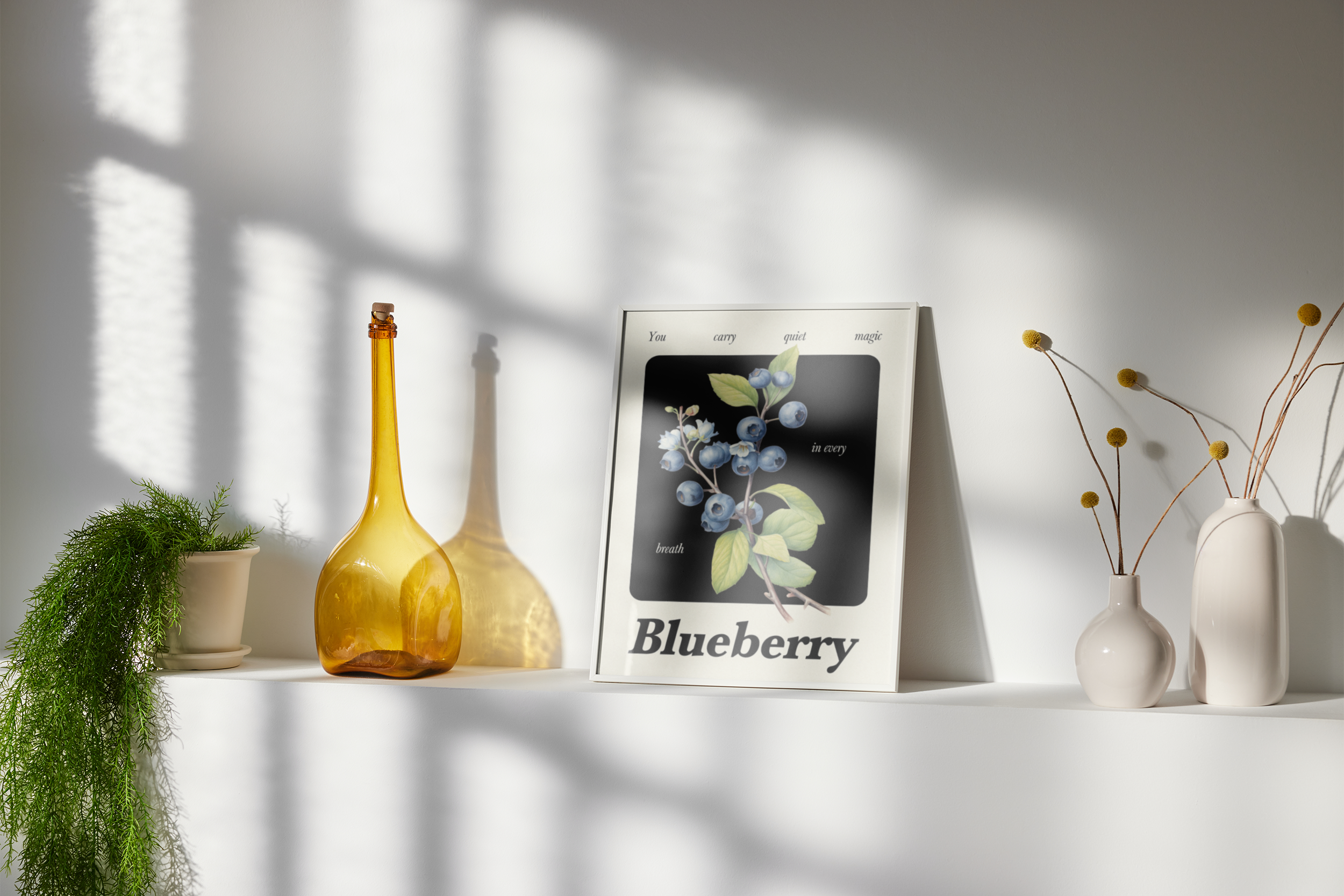 A small framed print featuring a dark image with a botanical illustration of blueberries on a branch and the word "Blueberry," propped on a white shelf next to a tall, amber glass bottle and two small vases with dry flowers.