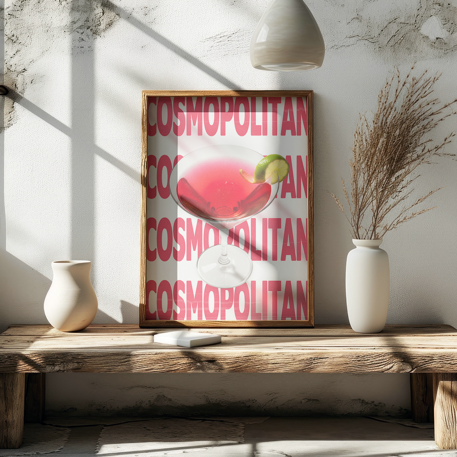 Framed poster of a pink Cosmopolitan cocktail with a lime garnish, against a background of repeating 'COSMOPOLITAN' text, resting on a rustic wooden bench in bright sunlight.