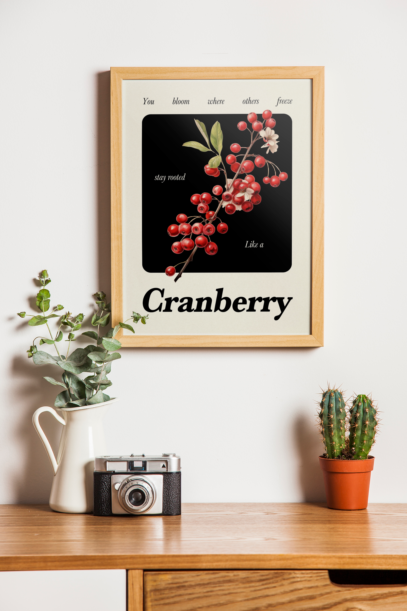 A wood-framed print featuring a botanical illustration of red cranberries on a vine against a black and cream background with short inspirational text, hanging above a wooden cabinet with a white pitcher, a vintage camera, and a potted cactus.