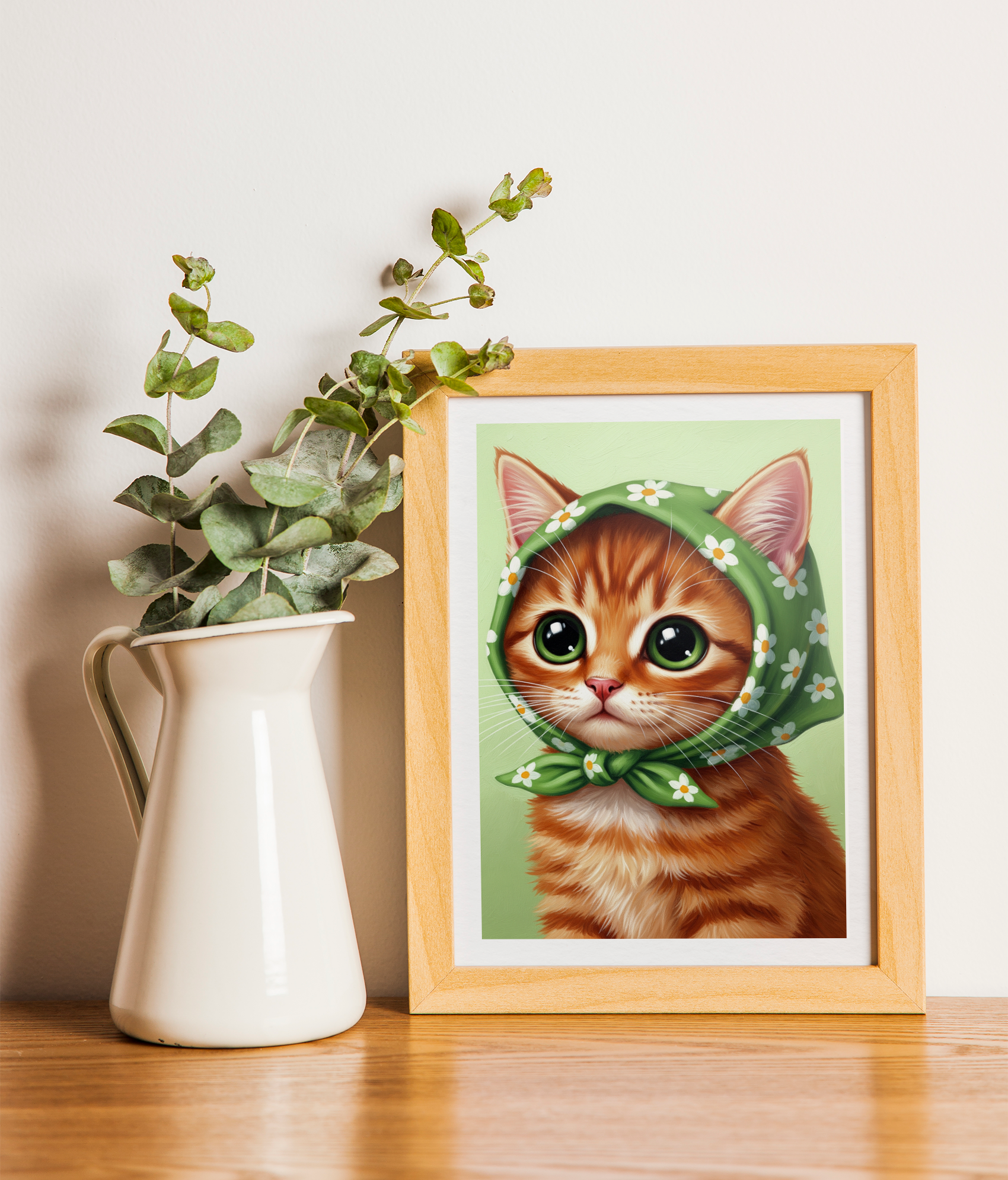 A wood-framed print of a close-up illustration of a ginger kitten with large eyes, wearing a green kerchief with white flowers, resting on a wooden surface next to a white pitcher holding eucalyptus branches.