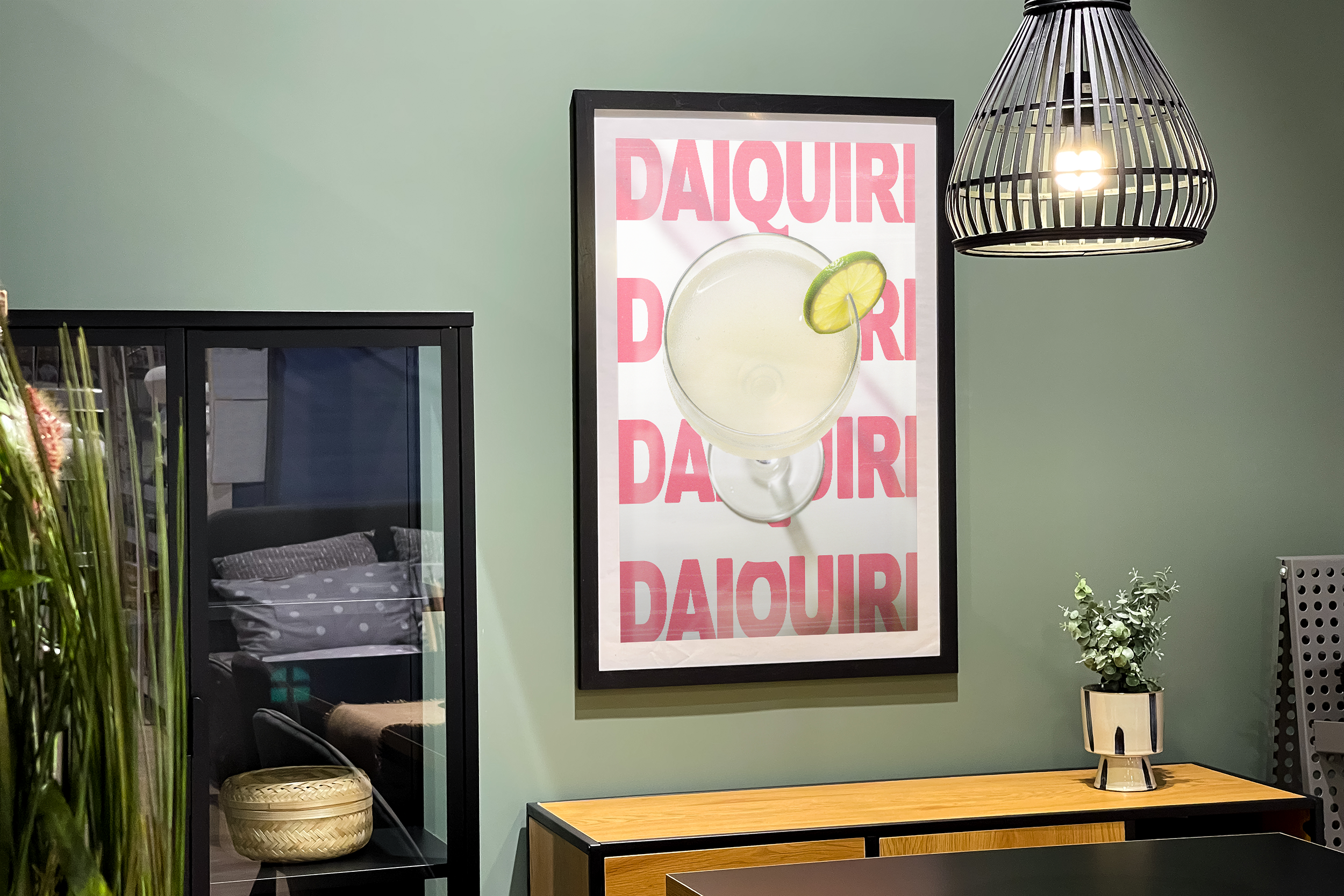 Black framed poster of a Daiquiri cocktail with a lime wheel, against a repeating pink and white background, hanging on a sage green wall above a wooden cabinet.