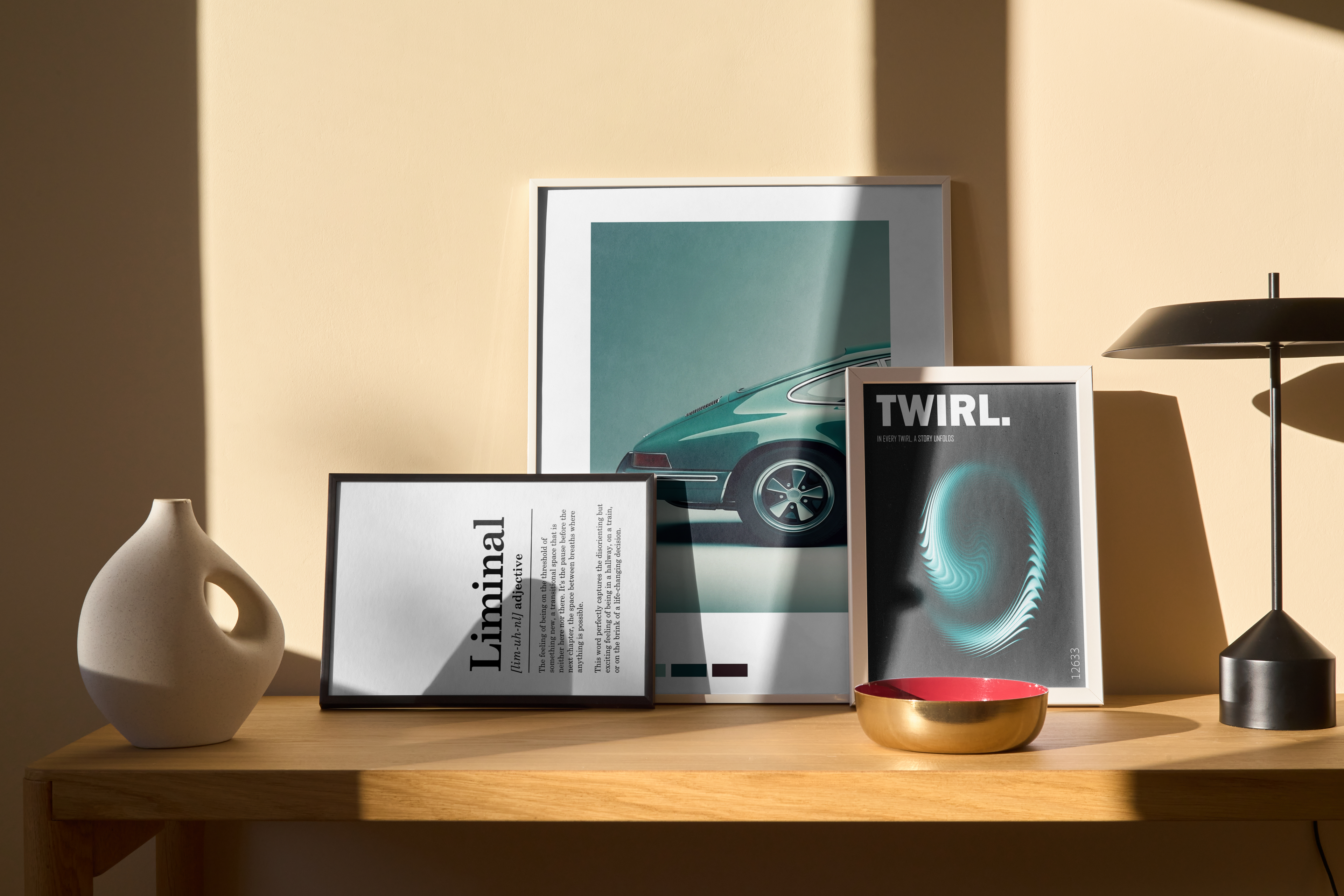 A group of three framed prints displayed on a wooden table: a dictionary definition of "Liminal," a modern abstract car poster, and a black-and-white "TWIRL" graphic, next to a black table lamp and a white vase.