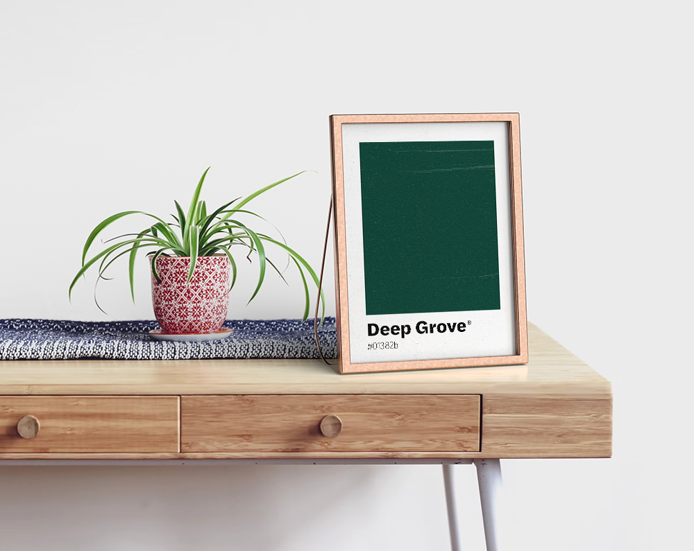 A simple yet elegant minimalist art print featuring a solid deep forest green color swatch labeled "Deep Grove" with its corresponding color code. Excellent for modern office decor, a creative studio, or for color enthusiasts. The framed print stands on a light wooden desk next to a vibrant spider plant in a patterned red and white pot, creating a focused and calming workspace.