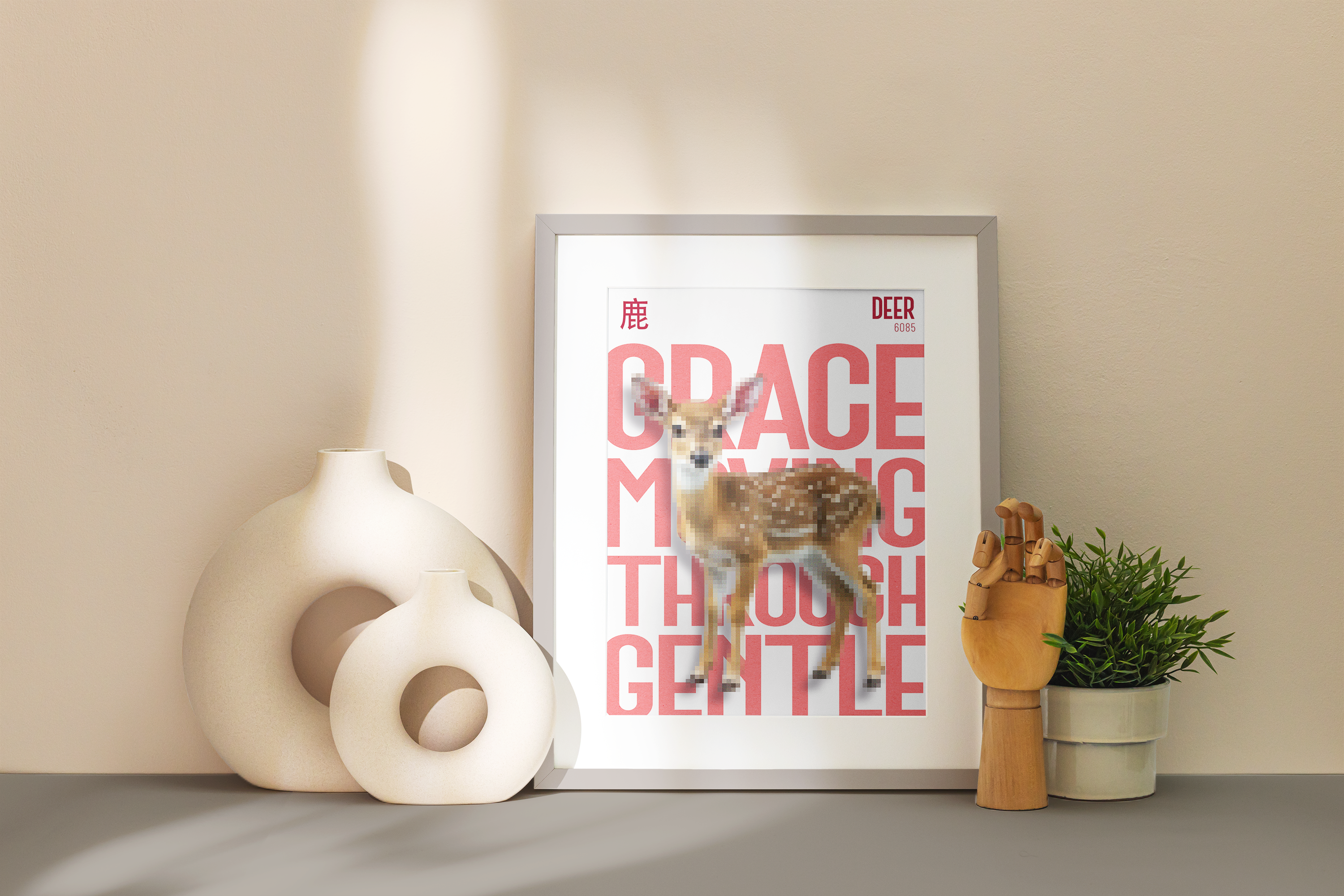 A framed poster of a small, spotted Deer (fawn), illustrated with a pixelated effect, sitting on a dark grey surface against a pale beige wall. Large pink text reads: "GRACE MOVING THROUGH GENTLE." The poster is flanked by abstract, circular white ceramic vases and a wooden jointed hand.