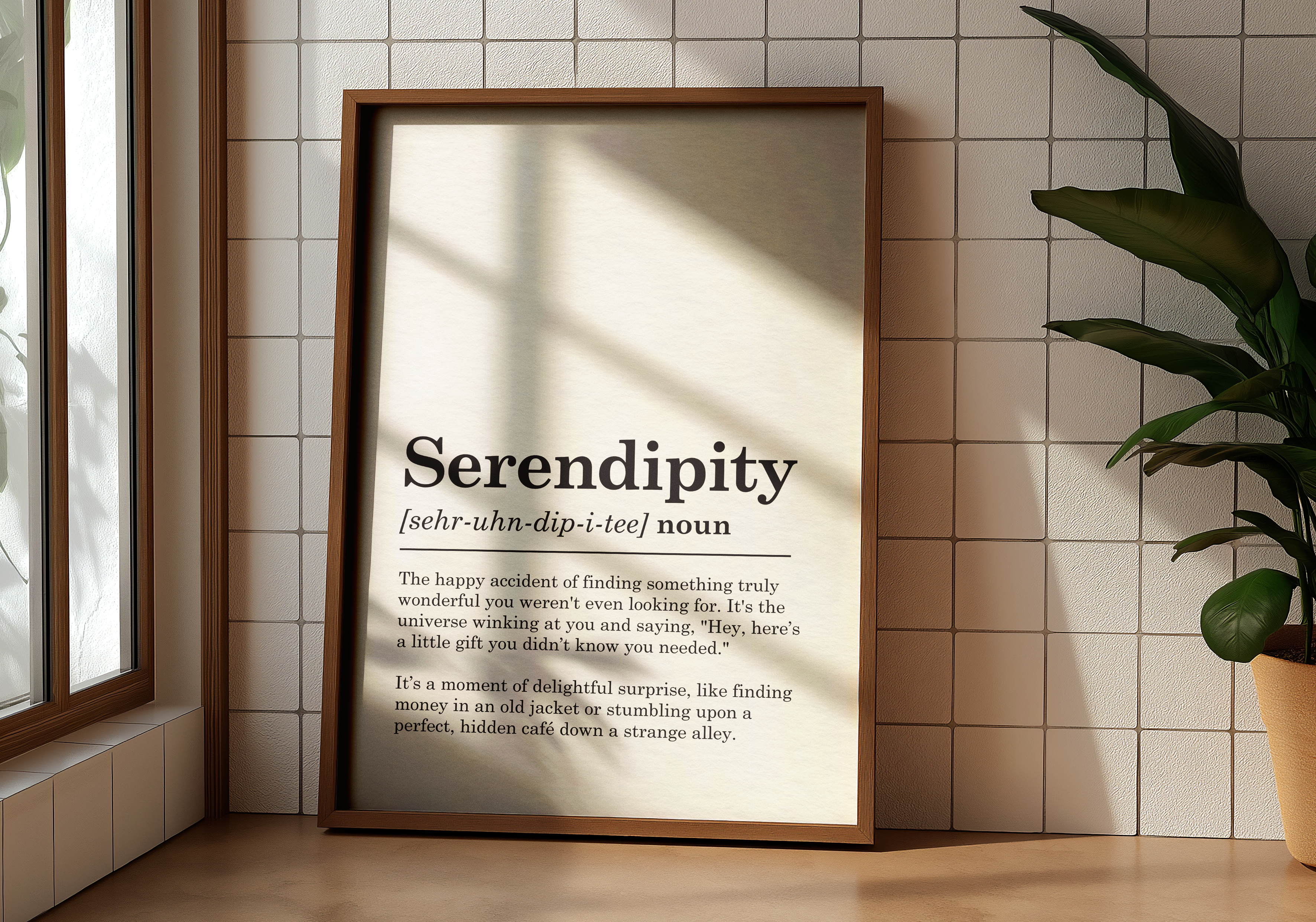 A large, brown-framed vertical print featuring the definition of the word 'Serendipity' leans against a white tiled wall. The print displays the word 'Serendipity' in a large, elegant font at the top, followed by its phonetic pronunciation and 'noun.' Below, a detailed explanation of the word's meaning is provided in clear black text. Soft natural light casts shadows across the tiled wall from a window that is partially visible on the far left.