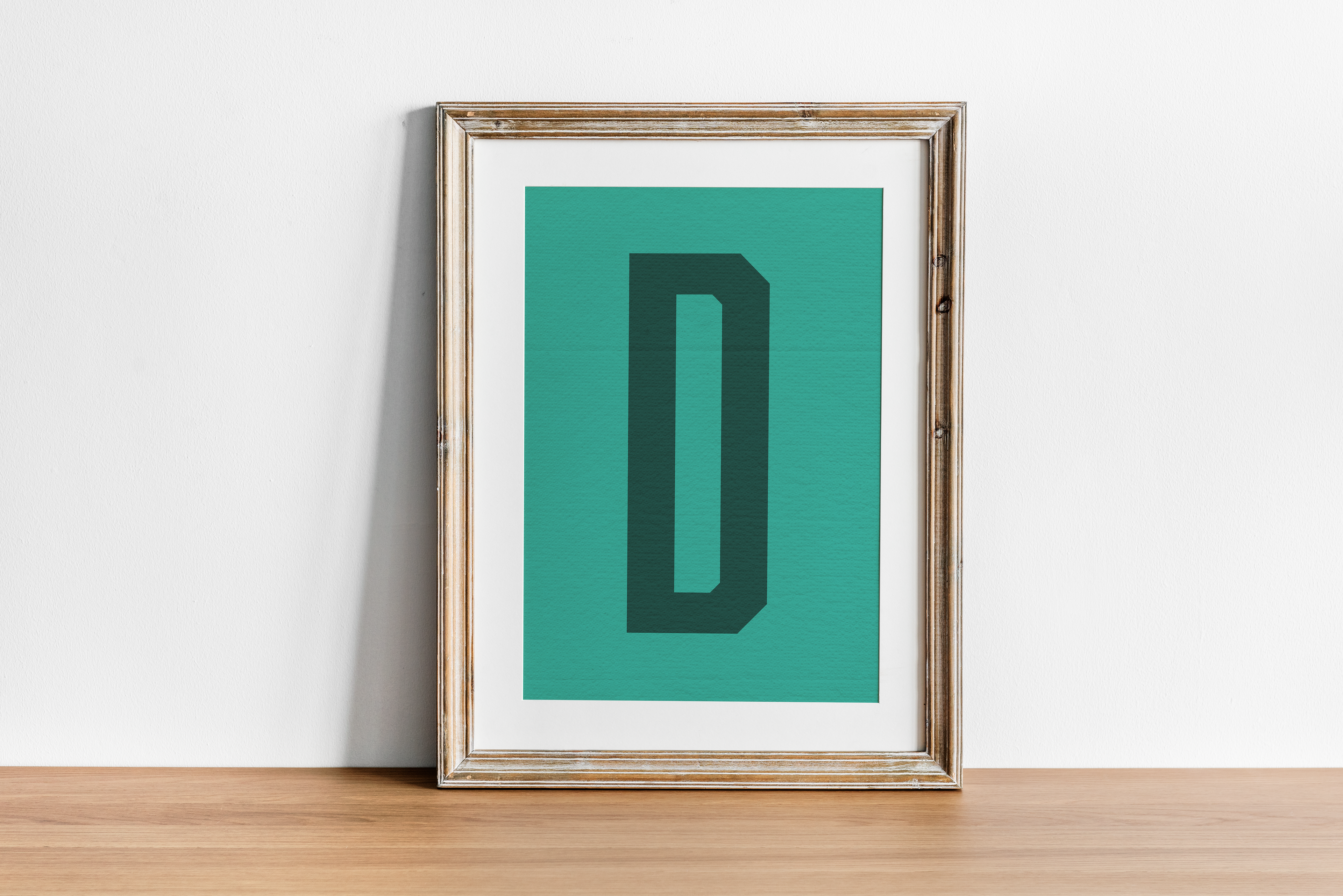 A framed print showcasing a dark green, 3D-style letter 'D' on a teal/turquoise background, resting on a wooden table against a clean white wall.