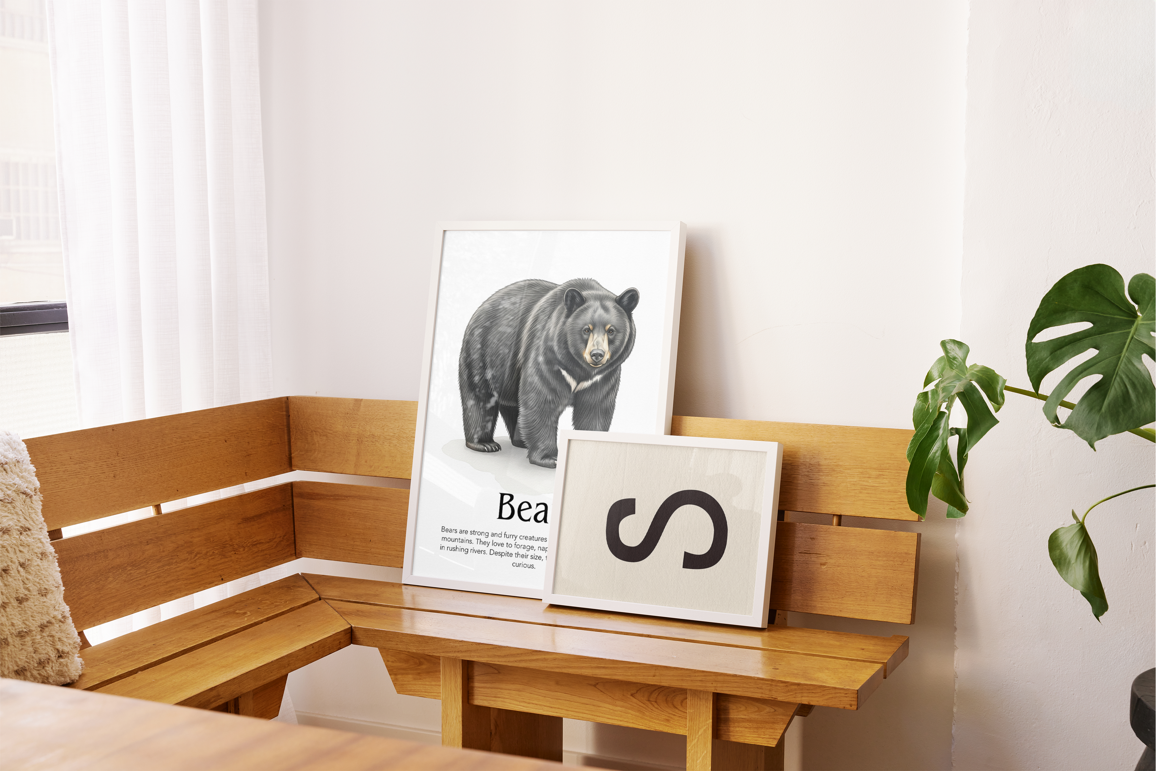 Two framed artworks rest on a light-colored wooden corner bench with a slatted back. The larger, vertical frame on the left features a detailed, realistic illustration of a black bear standing, with the word 'Bear' and descriptive text below it. To its right, a smaller, square frame displays a bold black letter 'S' on a cream-colored background. A large green plant with broad leaves is visible on the far right, and white curtains hang near a window on the left, letting in bright, soft daylight.