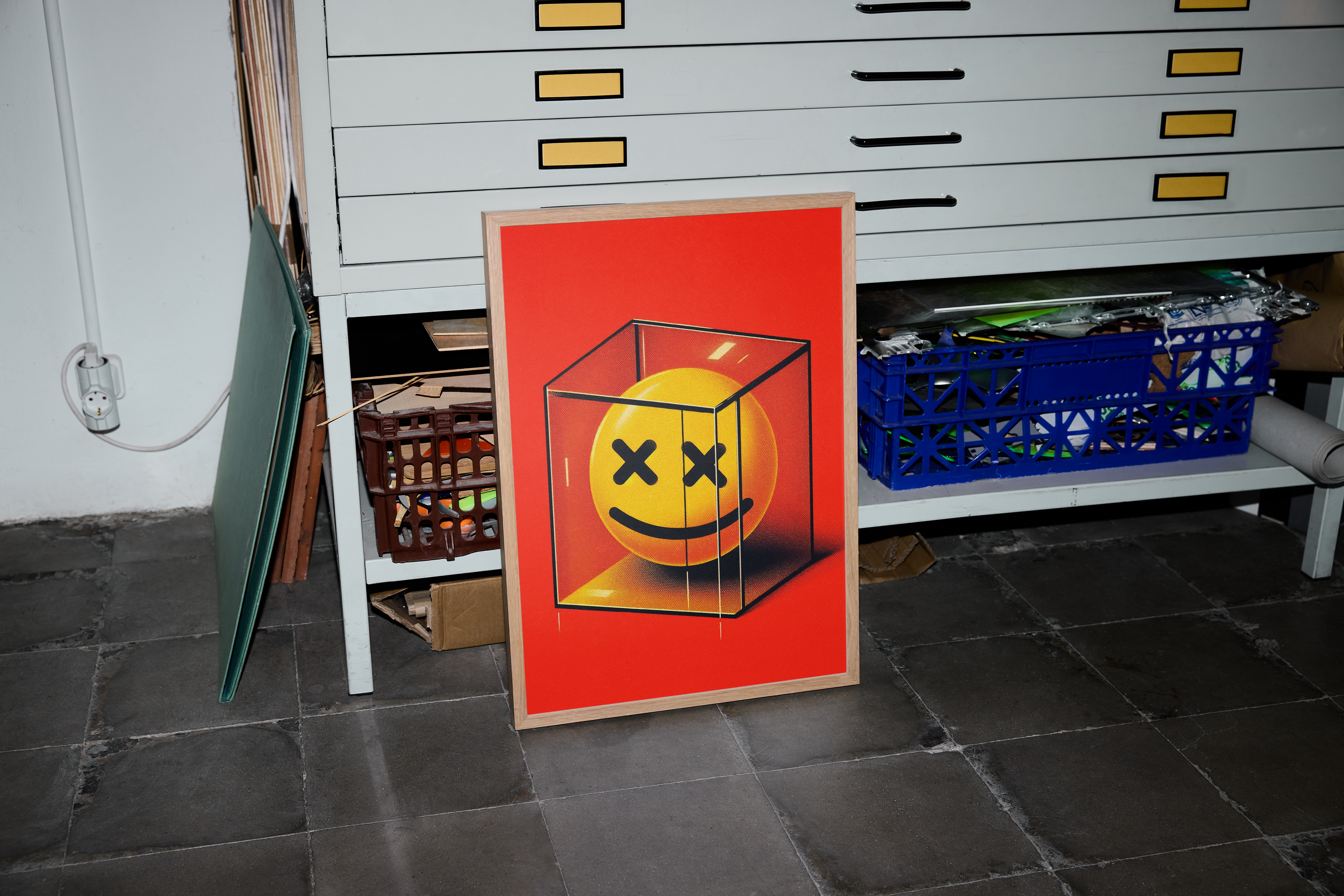 A colorful framed poster featuring a yellow smiley face inside a black wire cube, set against a vibrant red background. The artwork is leaning against a clean, white industrial metal shelving unit with yellow drawer fronts, suggesting a storage space or garage environment.