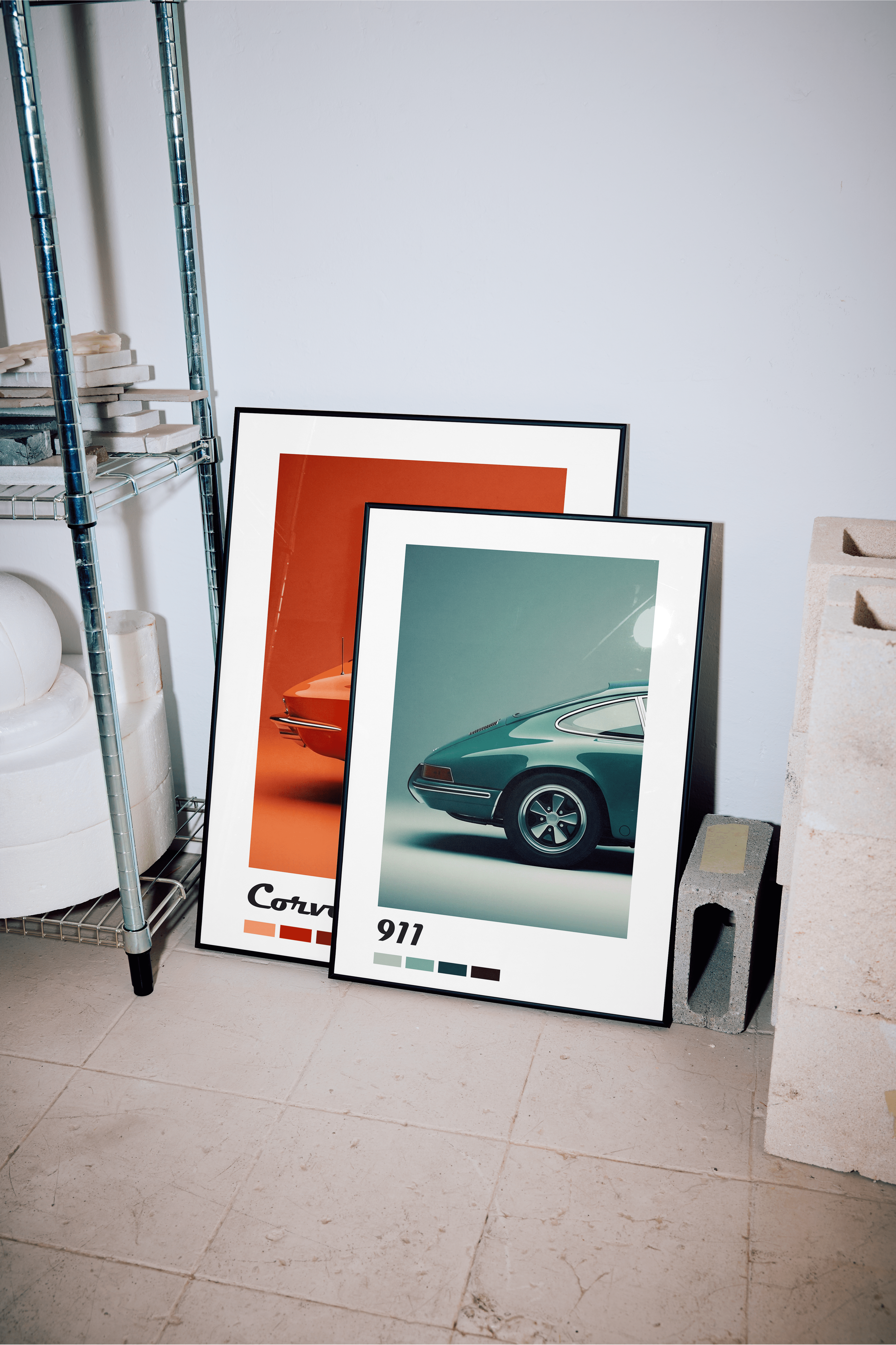 Two framed art prints of classic Porsche 911 cars lean against a light-colored wall in what appears to be a garage or workshop. The front print, slightly smaller, features a teal-colored Porsche with "911" and color swatches below it. Behind it, a larger print shows an orange-red Porsche with "Carrera" and color swatches at the bottom. To the left of the prints, a metal shelving unit holds various items, including white rolls or containers. To the right, several light grey concrete blocks are stacked. The f