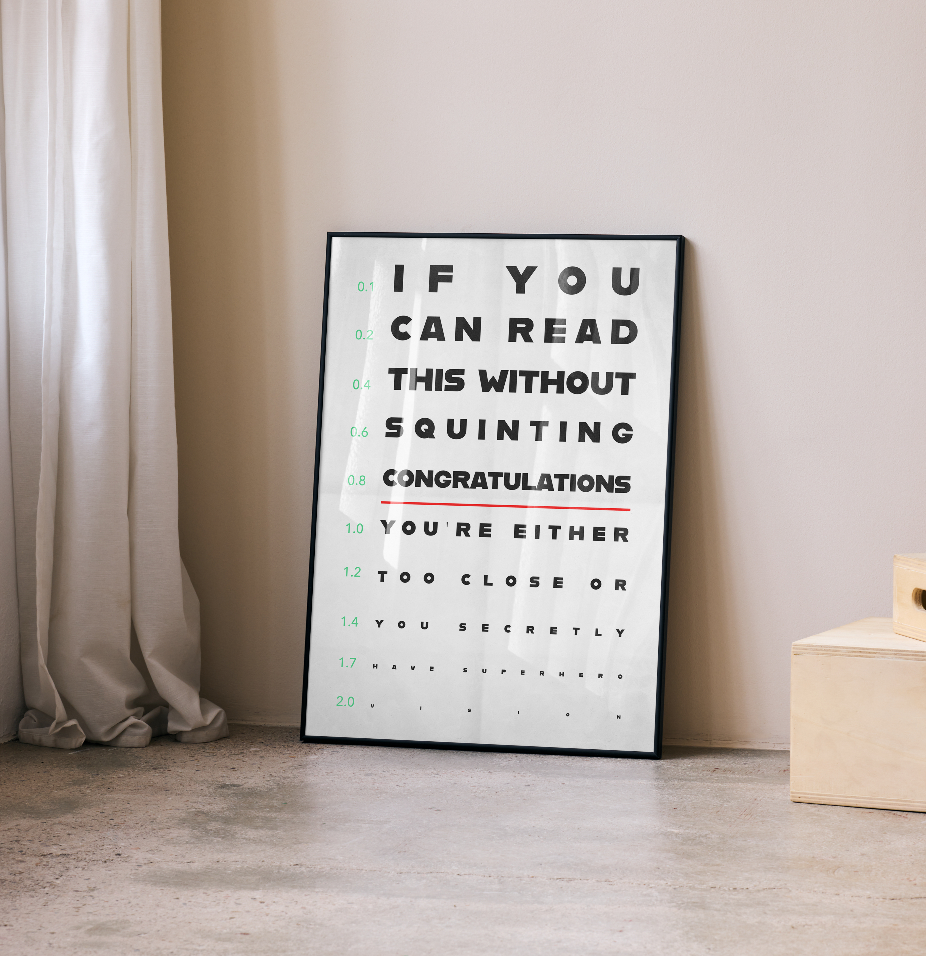 A black-framed poster designed as an eye chart with the humorous message "IF YOU CAN READ THIS WITHOUT SQUINTING CONGRATULATIONS YOU'RE EITHER TOO CLOSE OR YOU SECRETLY HAVE SUPERHERO VISION," leaning in a corner next to a white curtain.