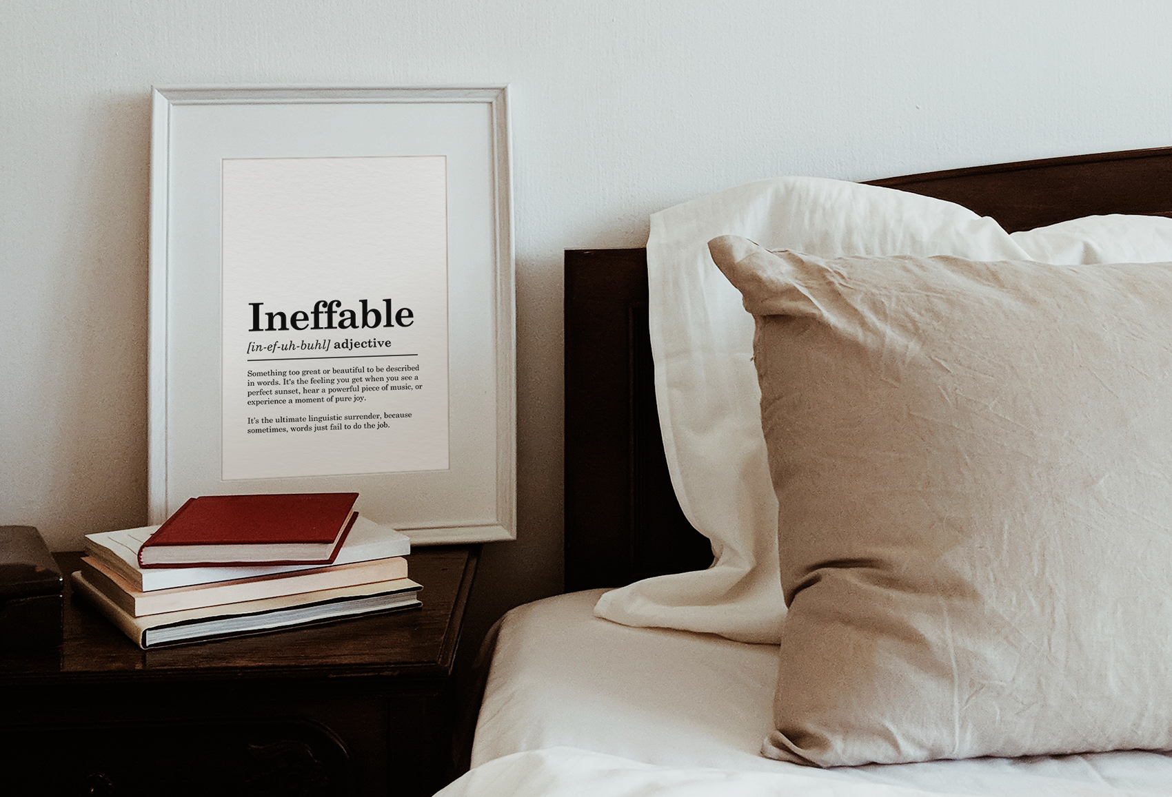 Framed print with the definition of the word 'Ineffable' on a bedside table next to a stack of books and a bed with white and beige pillows and bedding.