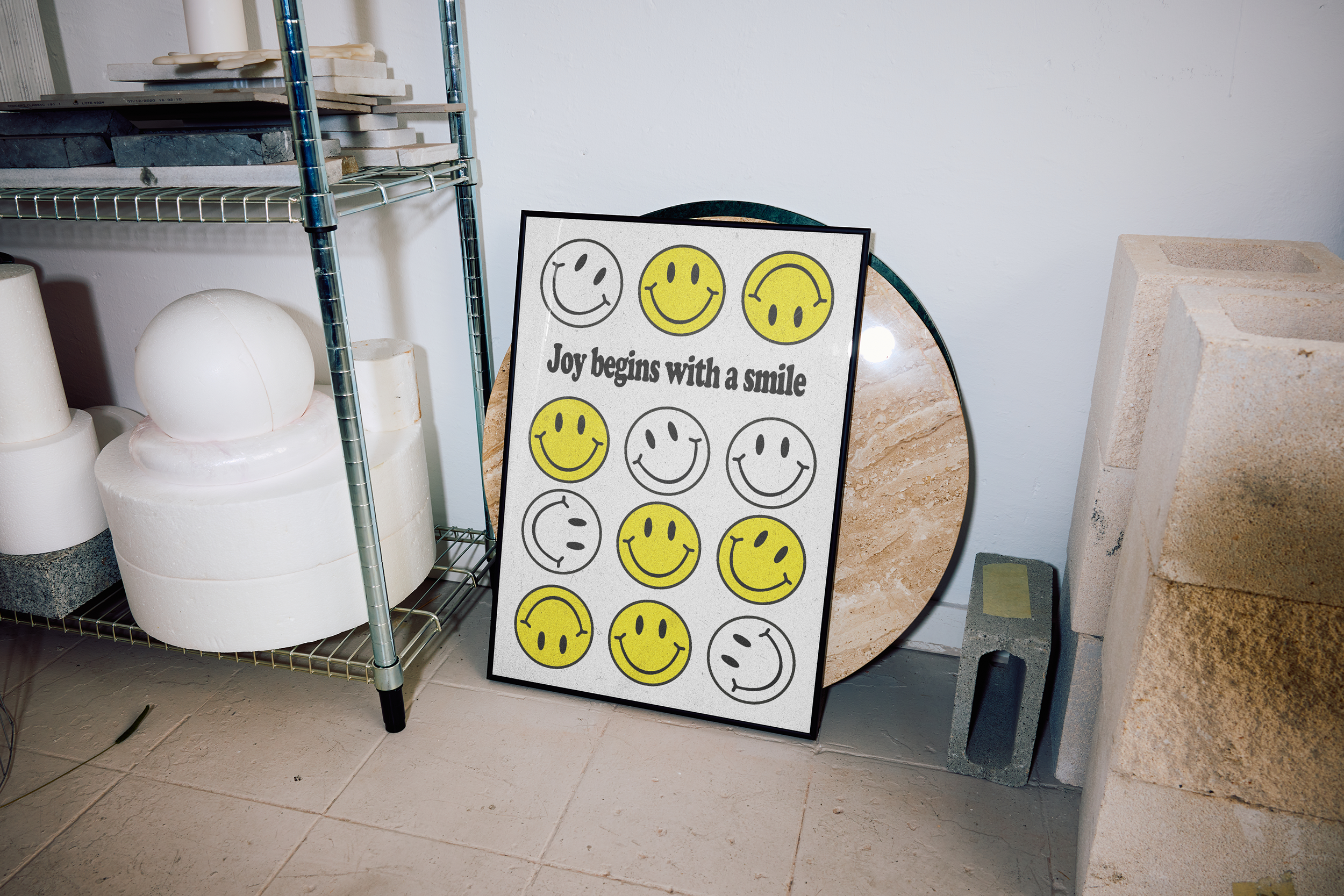 A framed poster featuring a grid of twelve smiley faces (some yellow, some outlined/partial) and the text "Joy begins with a smile," leaning against a wall in a workshop setting next to a marble-look table top and industrial shelving.