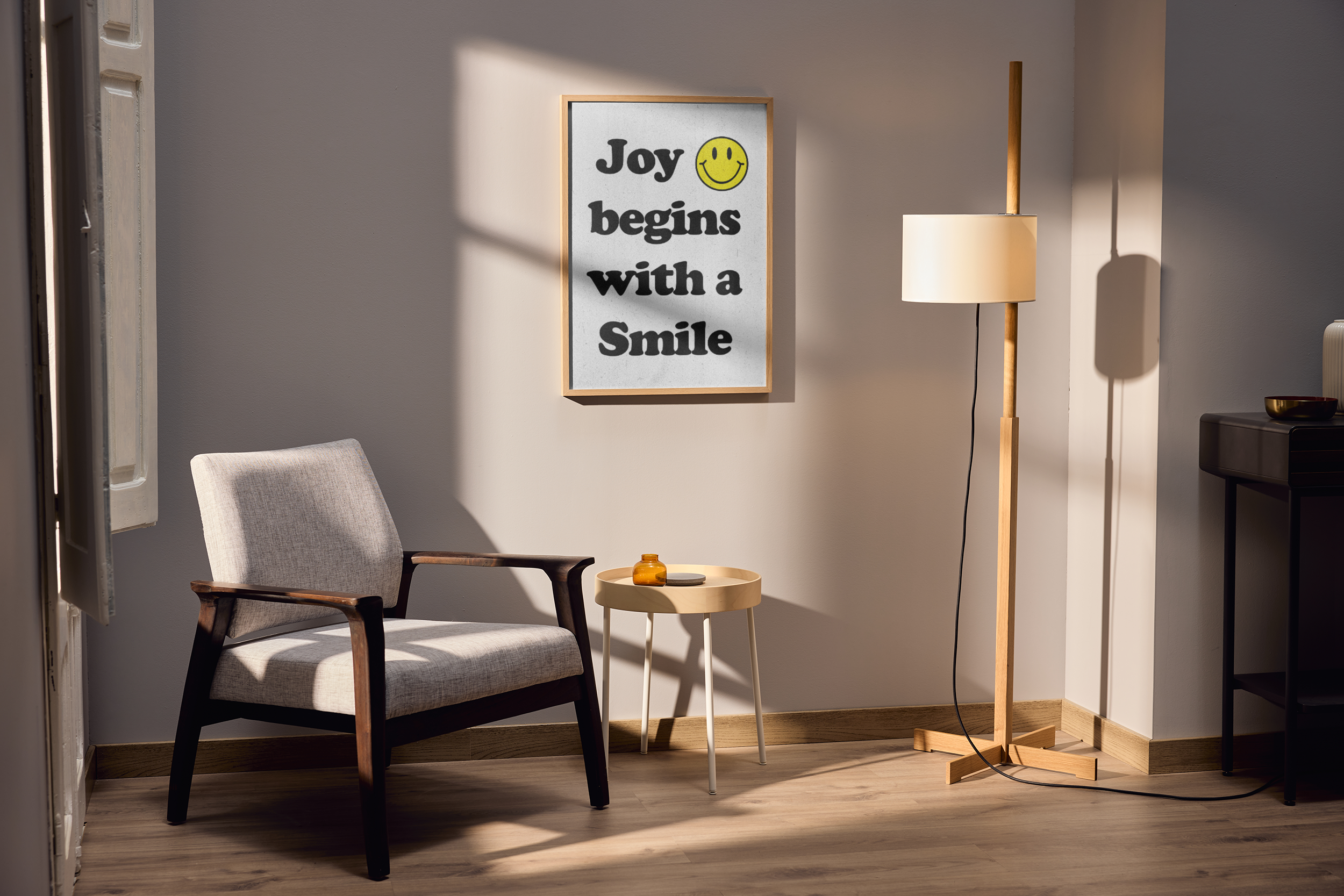 A wood-framed poster featuring the text "Joy begins with a Smile" with a small yellow smiley face icon, hanging on a light gray wall above a modern gray armchair and small side table.