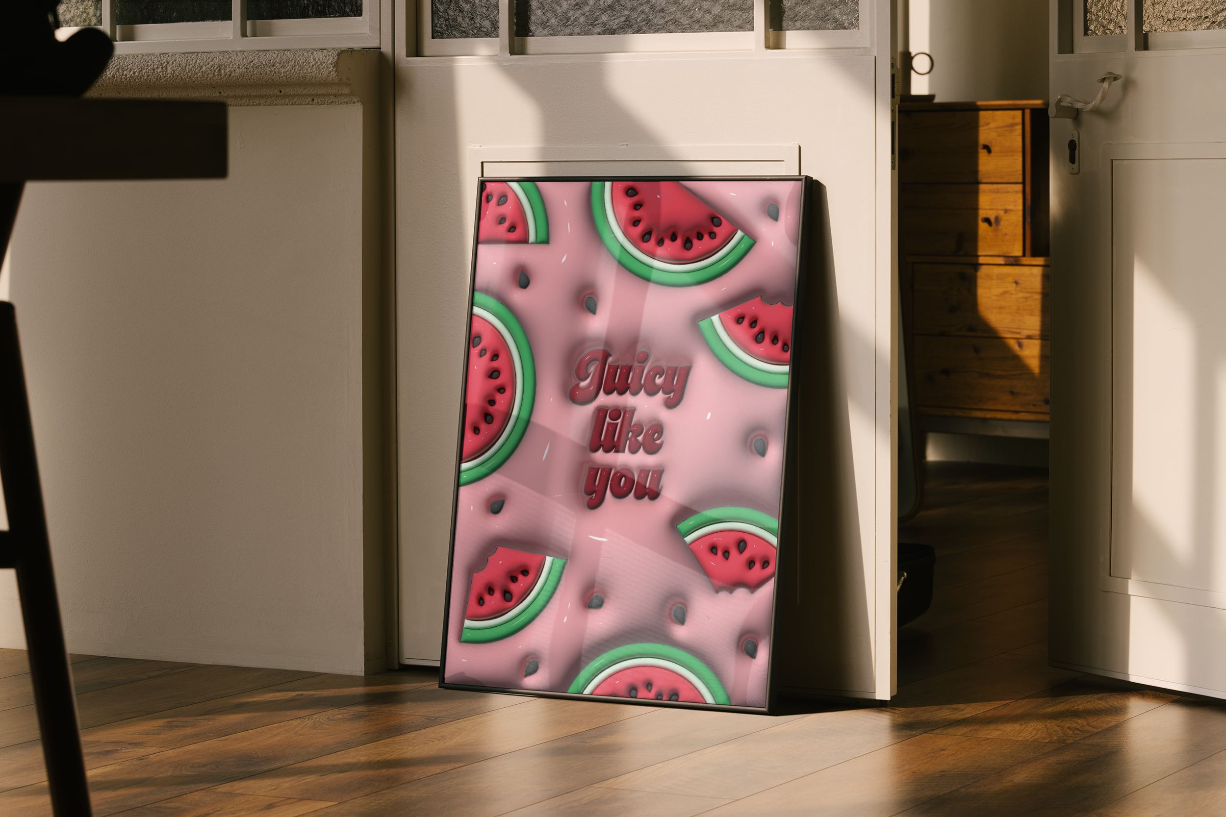 A black-framed poster featuring a pattern of 3D-rendered watermelon slices and the glossy pink text "Juicy like you," leaning against an open door frame on a wooden floor in a hallway.