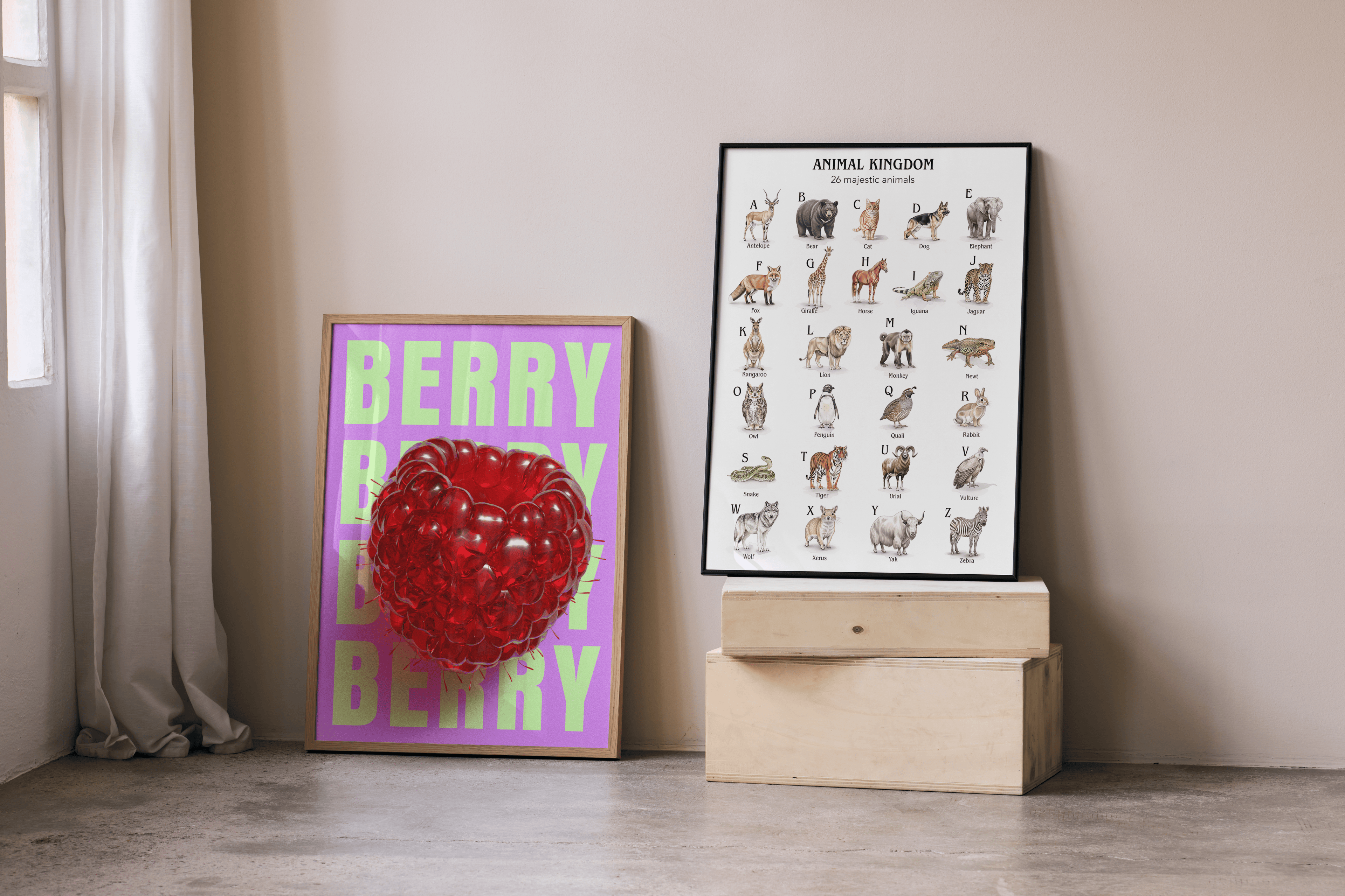 Two framed art prints lean against a light beige wall in what appears to be a kid's bedroom. On the left, a vibrant poster features a large, realistic 3D red raspberry centered on a purple background with the word "BERRY" in lime green text repeated above and below it. To the right, a larger framed poster titled "ANIMAL KINGDOM" displays various animals, each corresponding to a letter of the alphabet, on a white background. This poster rests on two stacked light wooden crates. To the far left, a window with