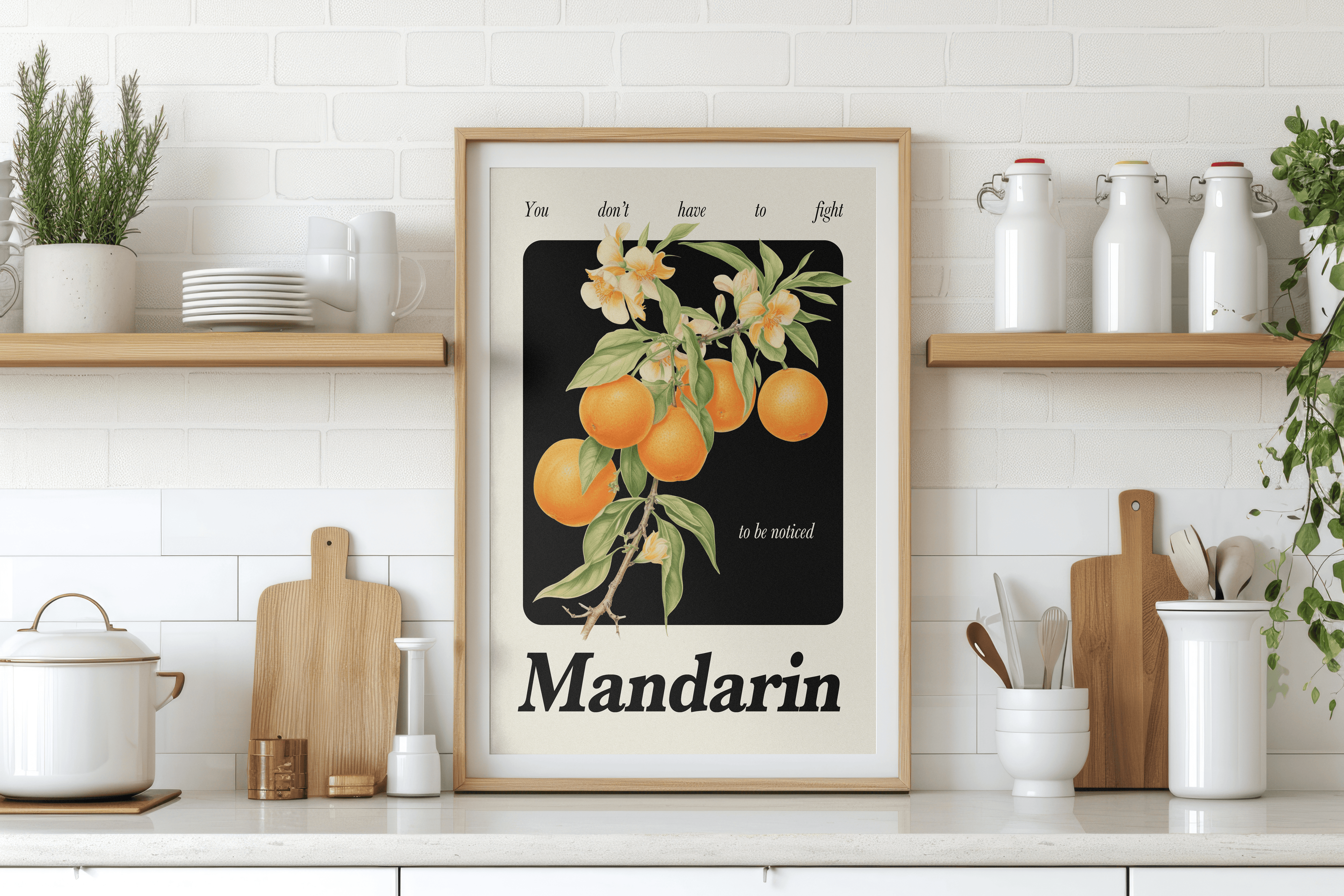 A framed art print featuring a cluster of mandarins on a branch with flowers, set against a dark background, is prominently displayed on a white kitchen countertop. The print has "You don't have to fight to be noticed" at the top and "Mandarin" at the bottom. The background wall is composed of white subway tiles. On either side of the print, wooden shelves are mounted on the wall. The left shelf holds white dishes and a potted herb plant. The right shelf has white ceramic containers with swing-top lids and 