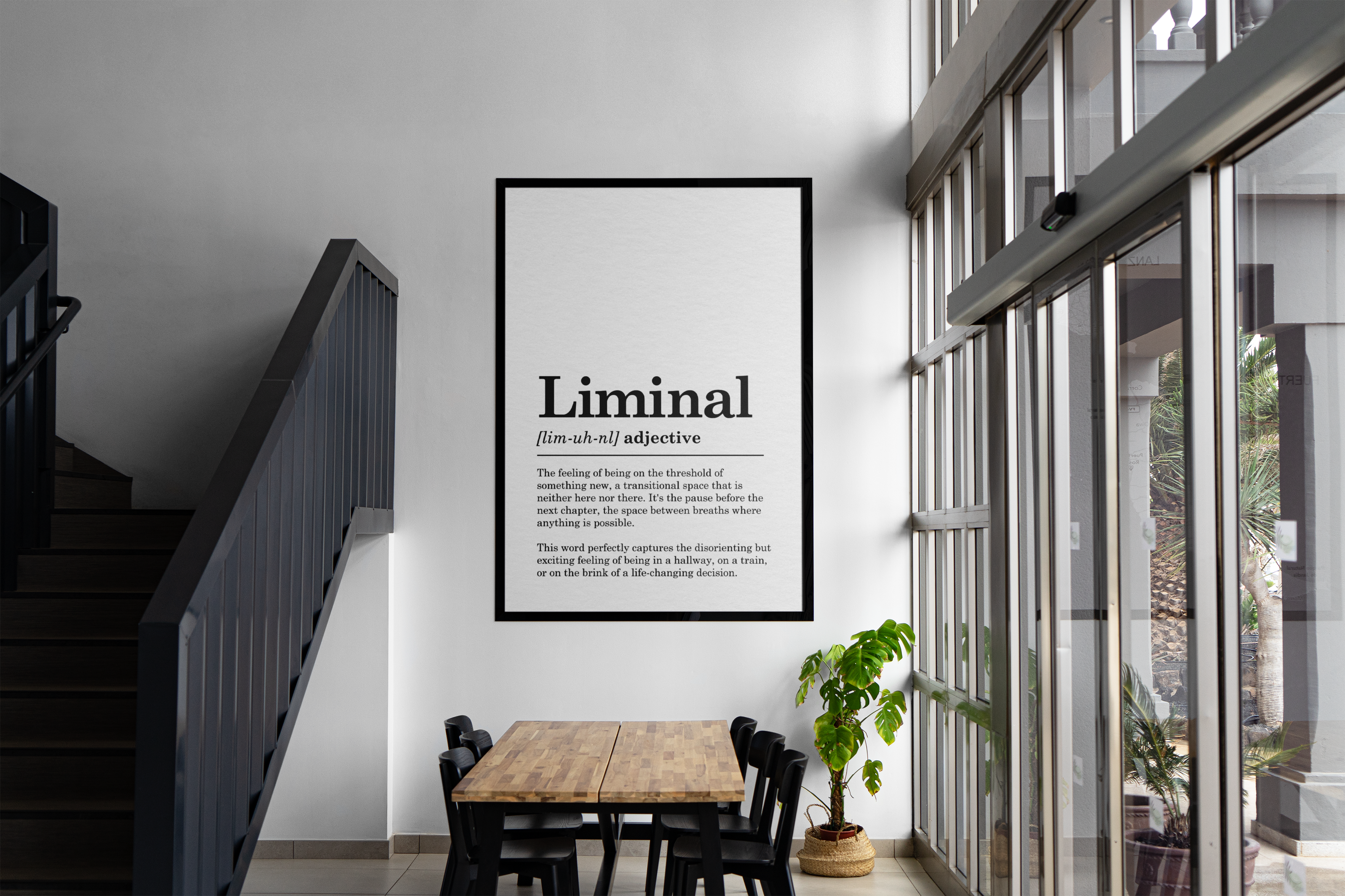 A huge, black-framed, vertical poster displaying the dictionary definition of the word 'Liminal' is centered on a stark white wall next to a large glass window wall. The text is clear and formal, detailing the word's meaning as 'the feeling of being on the threshold.' Below the art, a long wooden table surrounded by five black modern chairs is set up for dining or meetings. To the left, a dark-colored staircase with a black metal railing leads upwards.