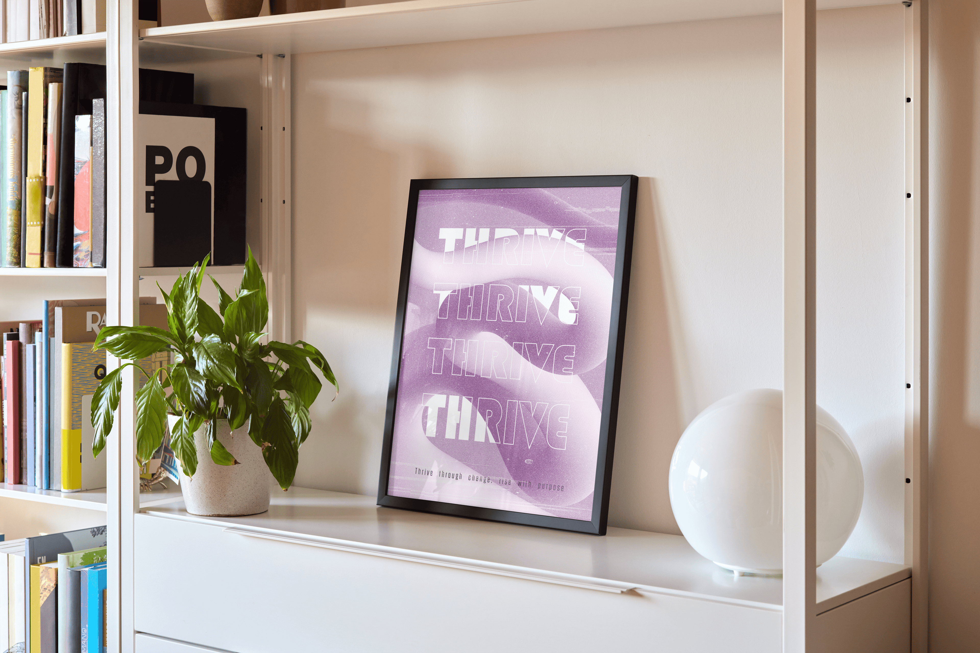 A framed art print is propped against a white wall on a modern white bookshelf unit. The print features the word "THRIVE" repeated three times in a white, wavy font on a textured purple background. To the left of the print, a potted green plant in a light-colored textured pot sits on the shelf. To the right, a white spherical lamp rests on the shelf. The bookshelf unit itself is filled with books on the left side, indicating a living room or study setting.