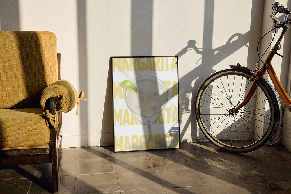 A fun and vibrant Pop Art print celebrating the classic Margarita cocktail, featuring a glass with a lime wedge set over a repeating yellow "MARGARITA" typography background. Perfect for a home bar, game room, or stylish corner. The framed print is leaning on a tiled floor, casting a striking shadow from the nearby vintage bicycle, next to a cozy mustard yellow armchair.