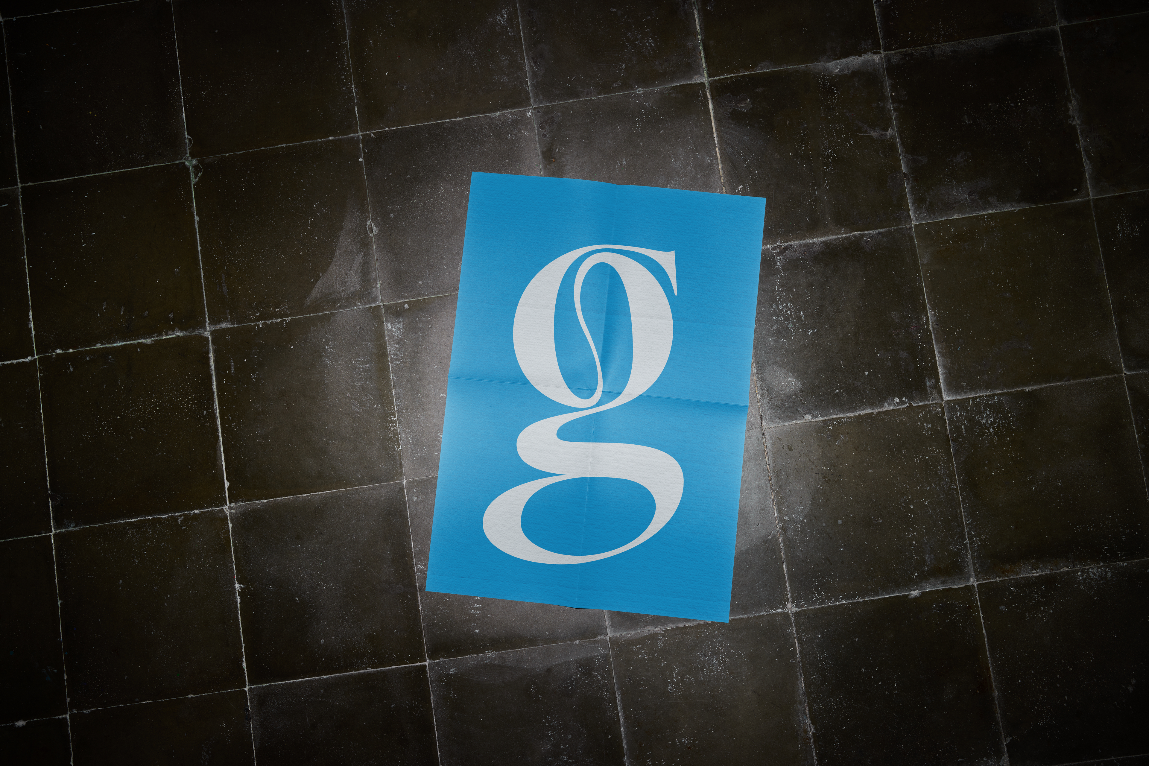 A mockup of a square poster design lying flat on a textured, dark gray tiled floor. The poster is a simple, minimalist design with a solid light blue background and a large, stylized white lowercase letter 'g' centered on the print.