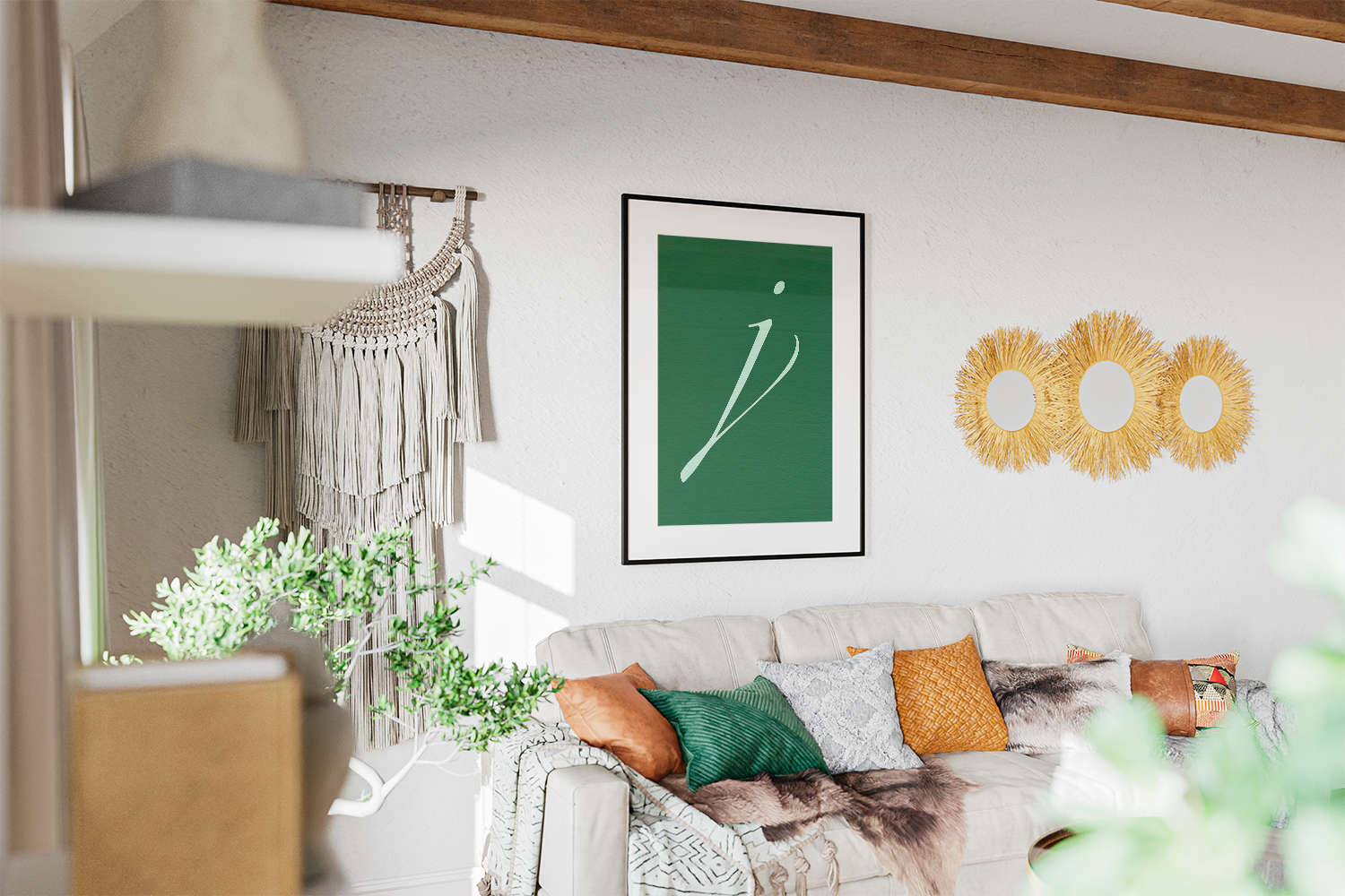 A stylish monogram art print featuring a clean, elegant white script letter 'J' on a vibrant emerald green background. Perfect for personalized home decor in a living room or bedroom. The framed print hangs above a neutral sofa, accented by cozy textured pillows (including green and orange), gold sunburst mirrors, and bohemian macrame wall hangings, creating a warm, textured, and eclectic space.