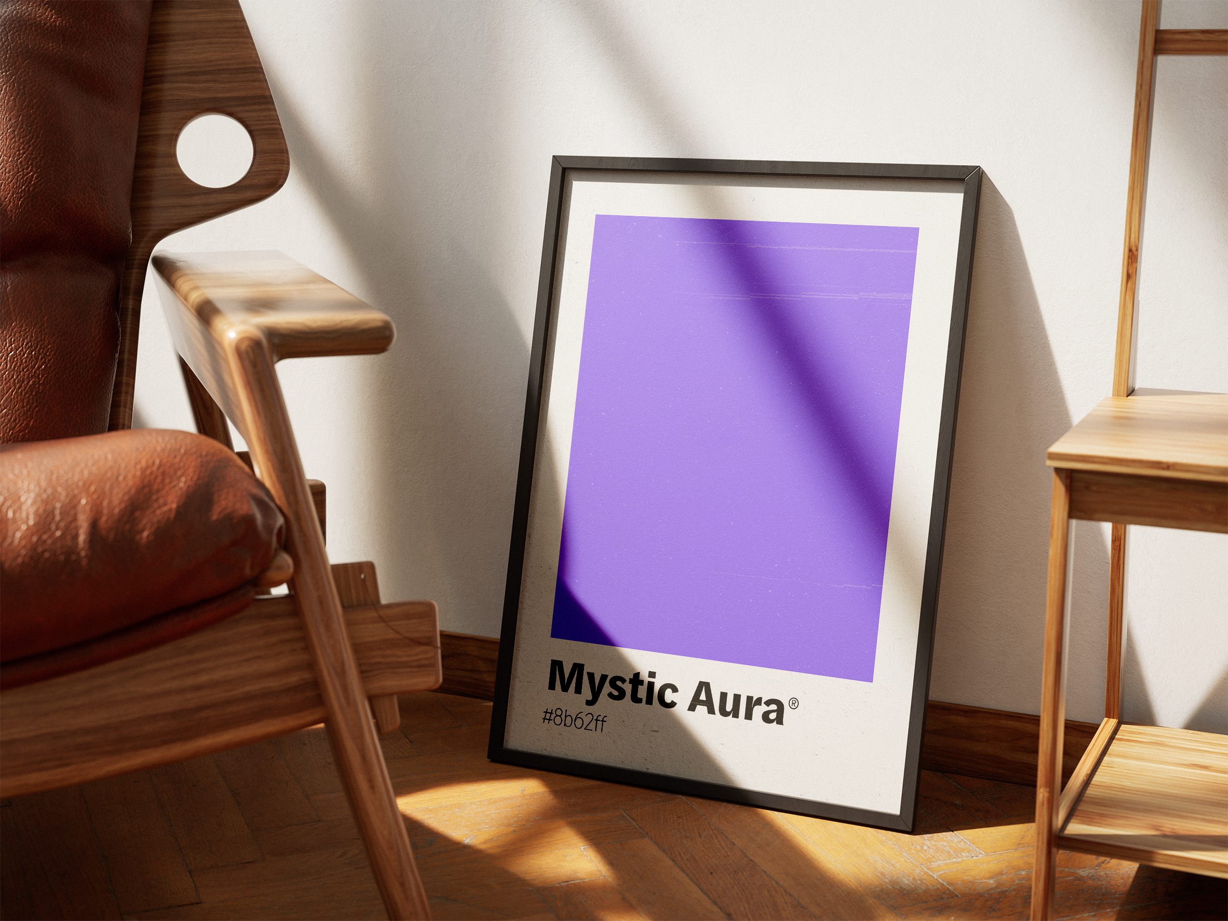 A black-framed print featuring a large block of purple labeled "Mystic Aura," leaning against a white wall on a wooden floor next to a leather and wood chair in a sunlit room.