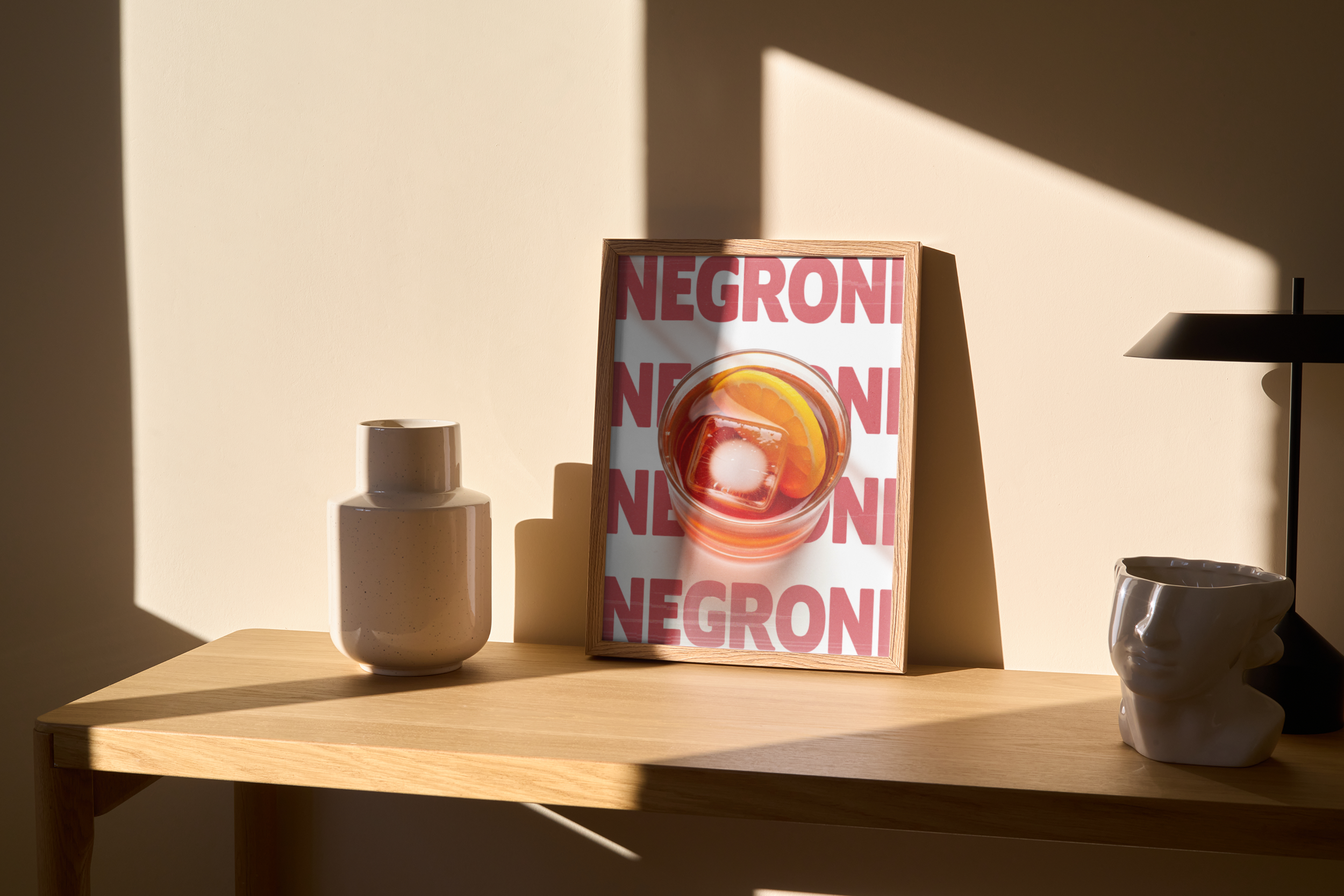 Small framed print of a Negroni cocktail with a slice of orange and ice, overlaid on a background of repeating 'NEGRONI' text, displayed on a light wooden table in warm sunlight.