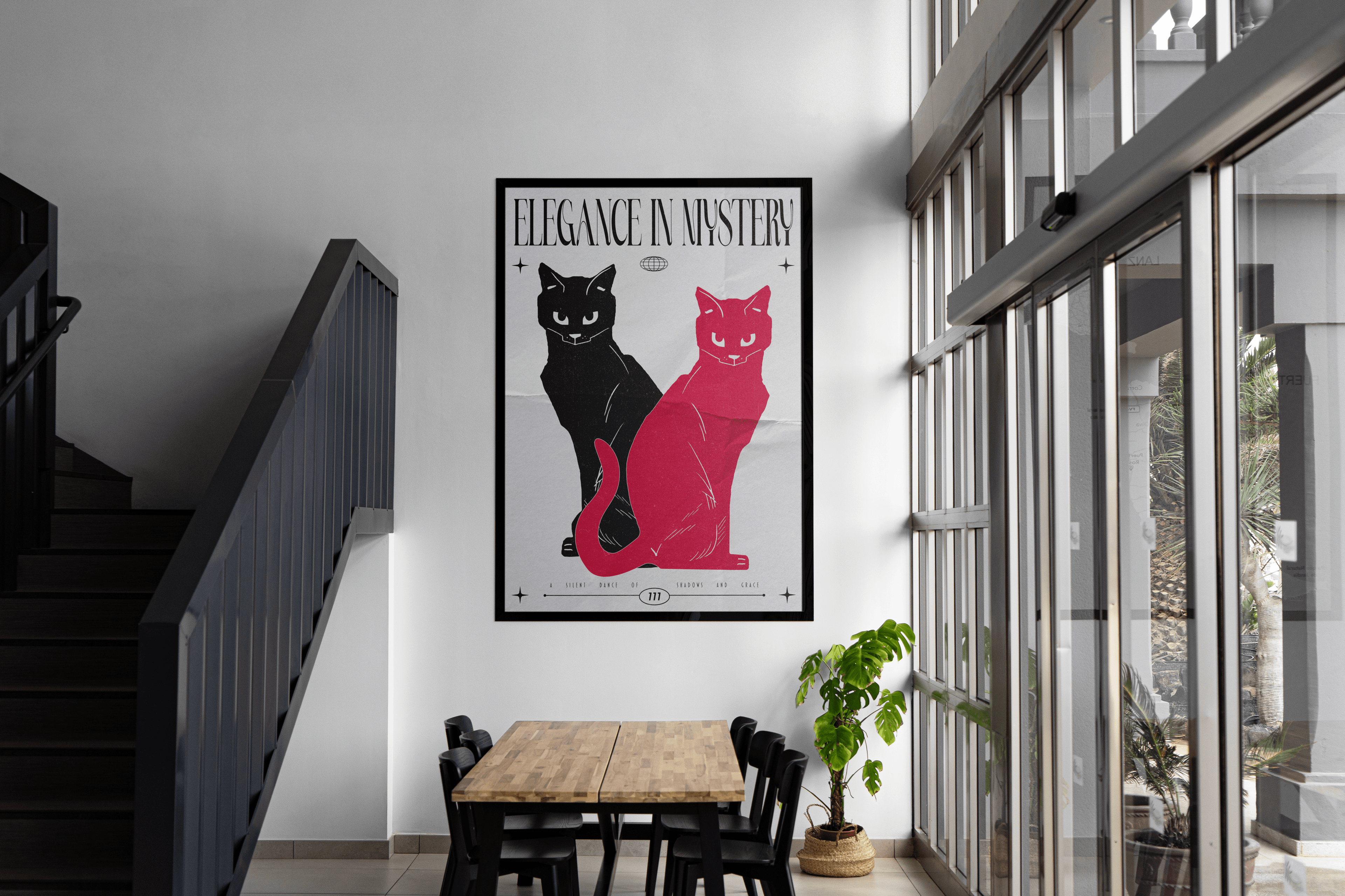 A large framed art print featuring a black cat and a pink cat sitting side-by-side, with the text "ELEGANCE IN MYSTERY" above them, is prominently displayed on a white wall. To the left of the print, a dark staircase with black railings ascends. Below the print, a light wooden table with two sections is surrounded by black chairs. To the right, a large window with multiple panes overlooks an outdoor area with plants. A small green plant in a woven pot sits near the table. The space appears to be a modern of