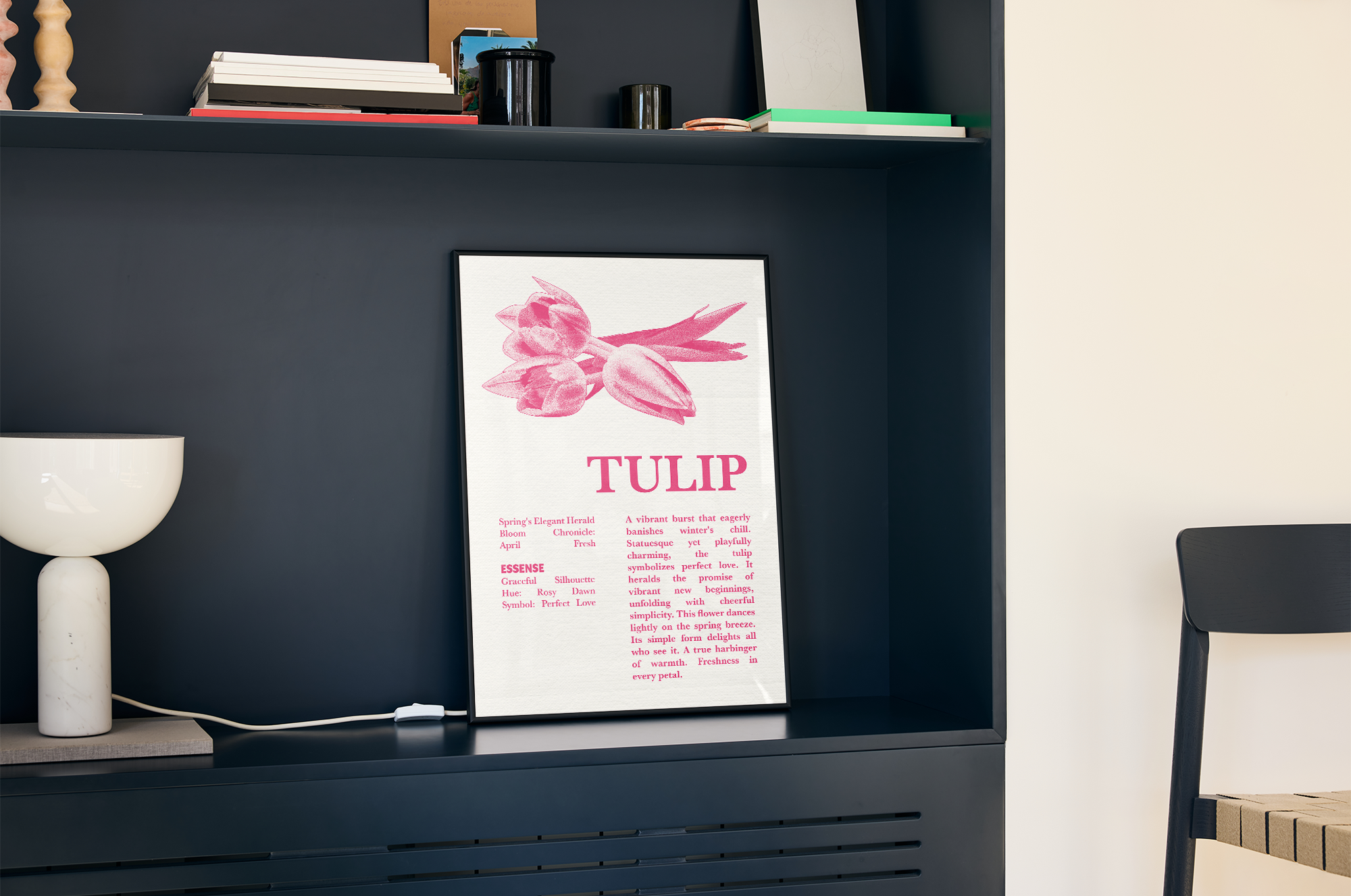 A framed print with a minimalist illustration of a pink tulip flower and the title "TULIP," alongside a detailed description and essential attributes, displayed inside a dark blue shelving unit next to a white table lamp and various stacked books.