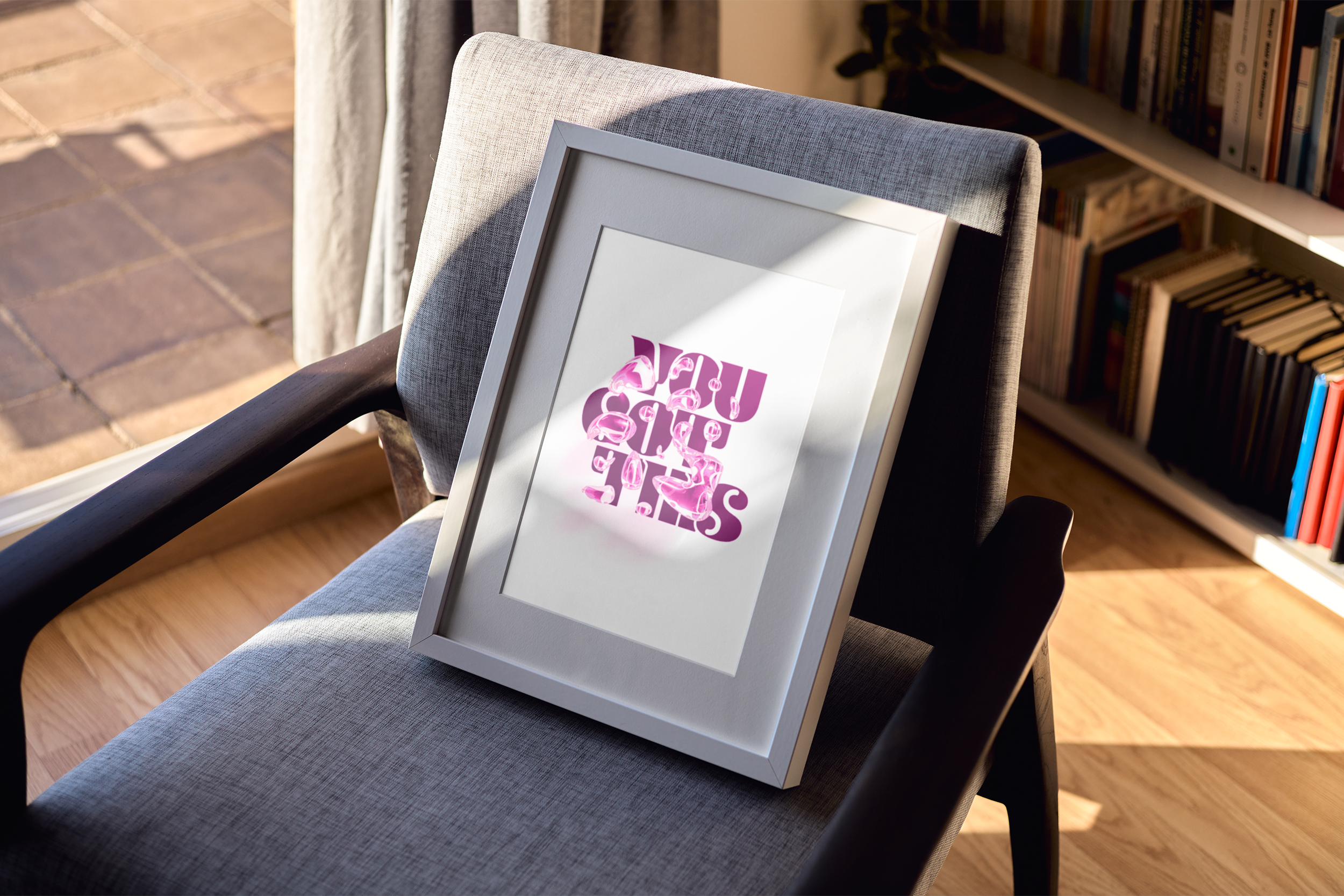 A gray-framed print with layered pink typography forming the phrase "YOU GOT THIS," resting on a gray armchair in a sunlit room with a full bookcase in the background.
