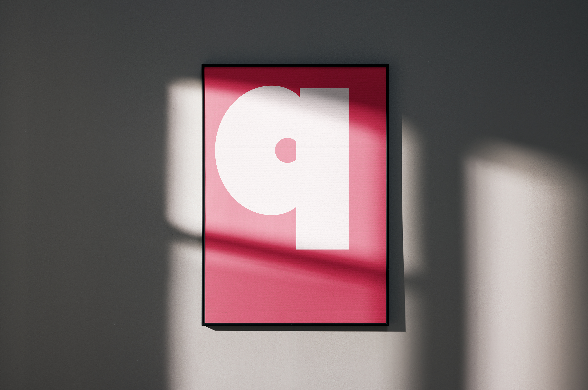 A black-framed poster with a large white graphic of a 'q' on a bright pink background, hanging on a grey wall. Strong diagonal sunlight casts shadows across the poster and wall.