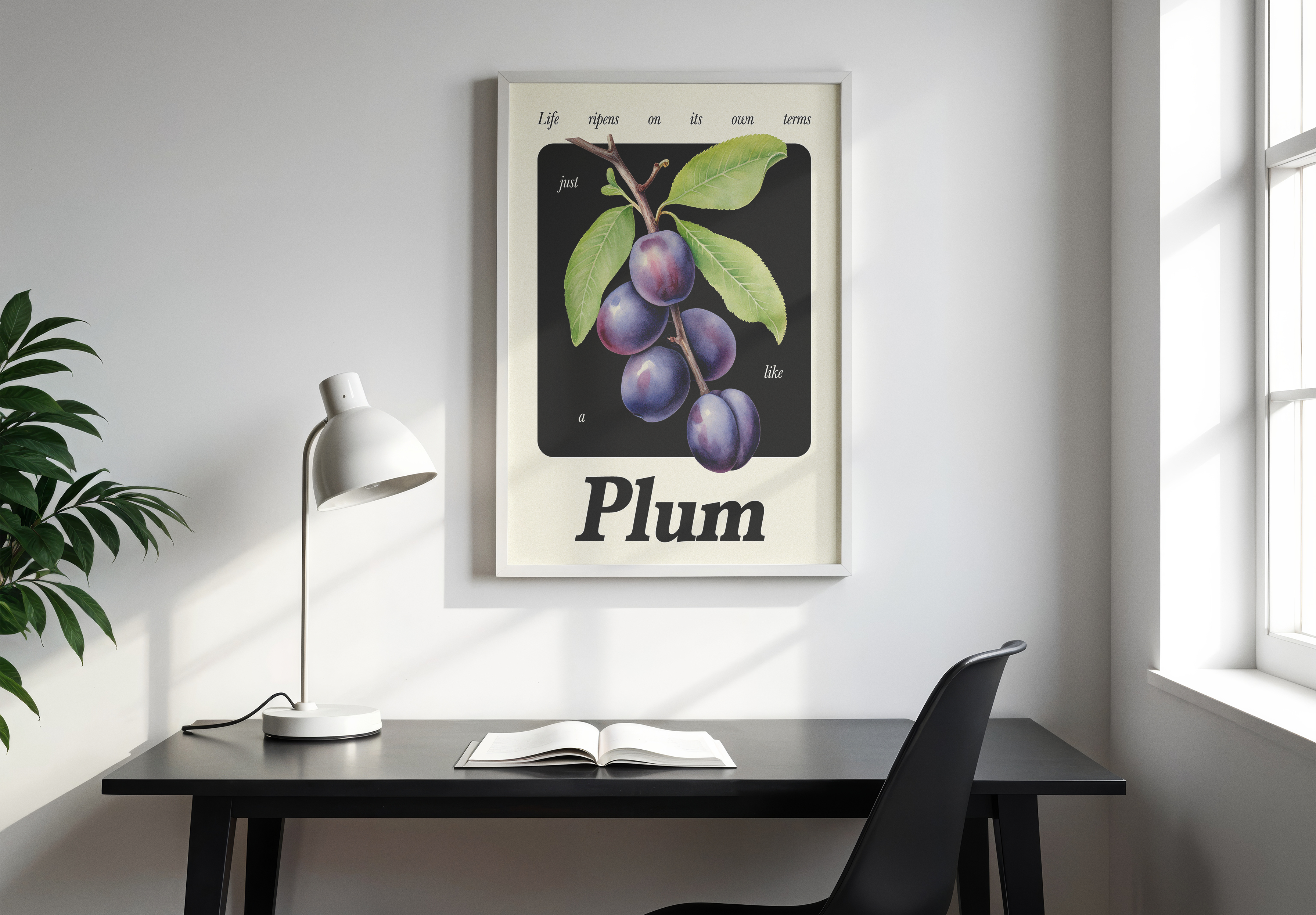 A white-framed botanical poster of Plums on a branch with an inspirational quote about life, hanging above a minimal black desk with a white lamp and an open book.