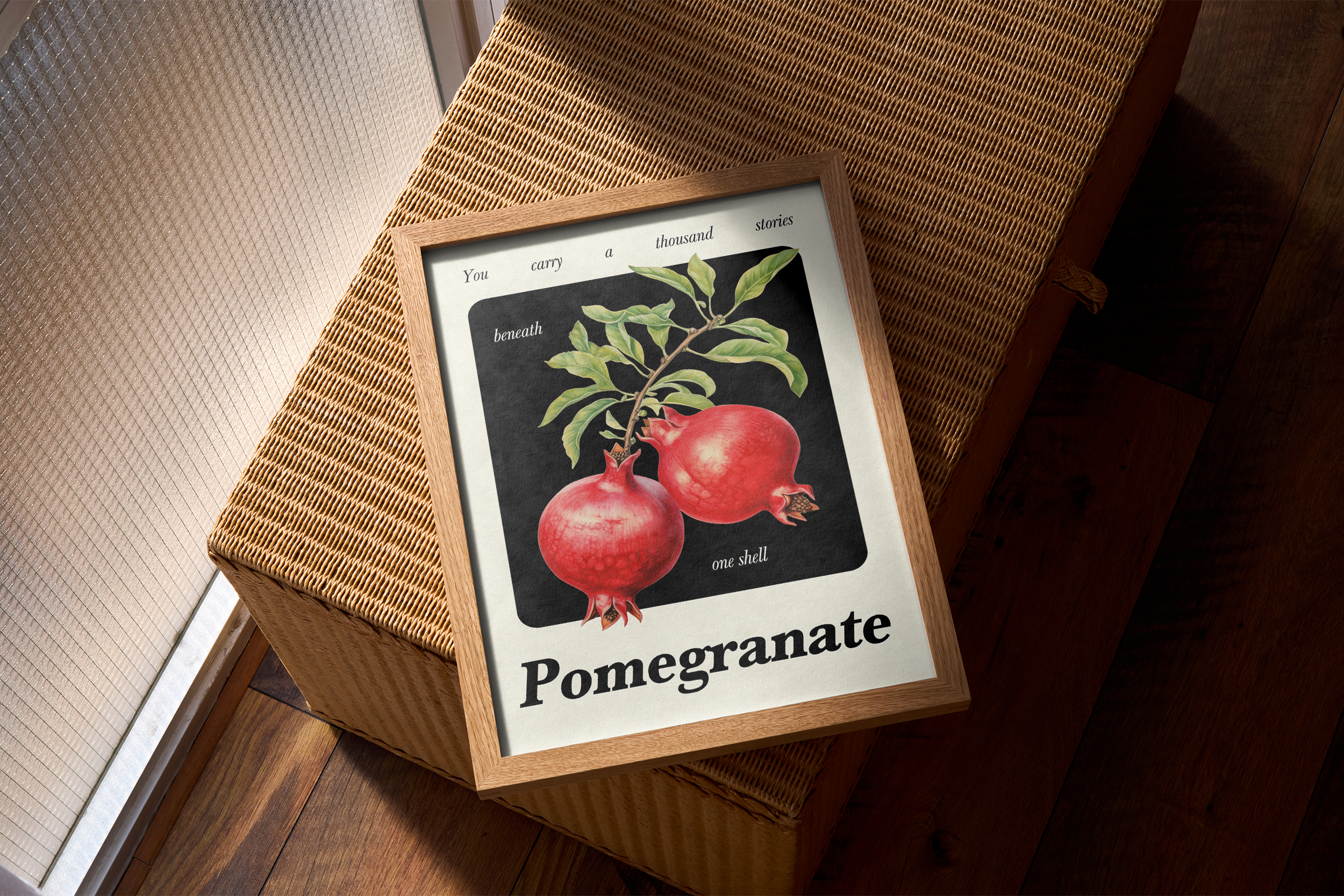 A wood-framed print featuring a botanical illustration of two pomegranates on a branch against a dark background, with the word "Pomegranate" and a surrounding quote, resting on a woven wicker chest or hamper next to a window.