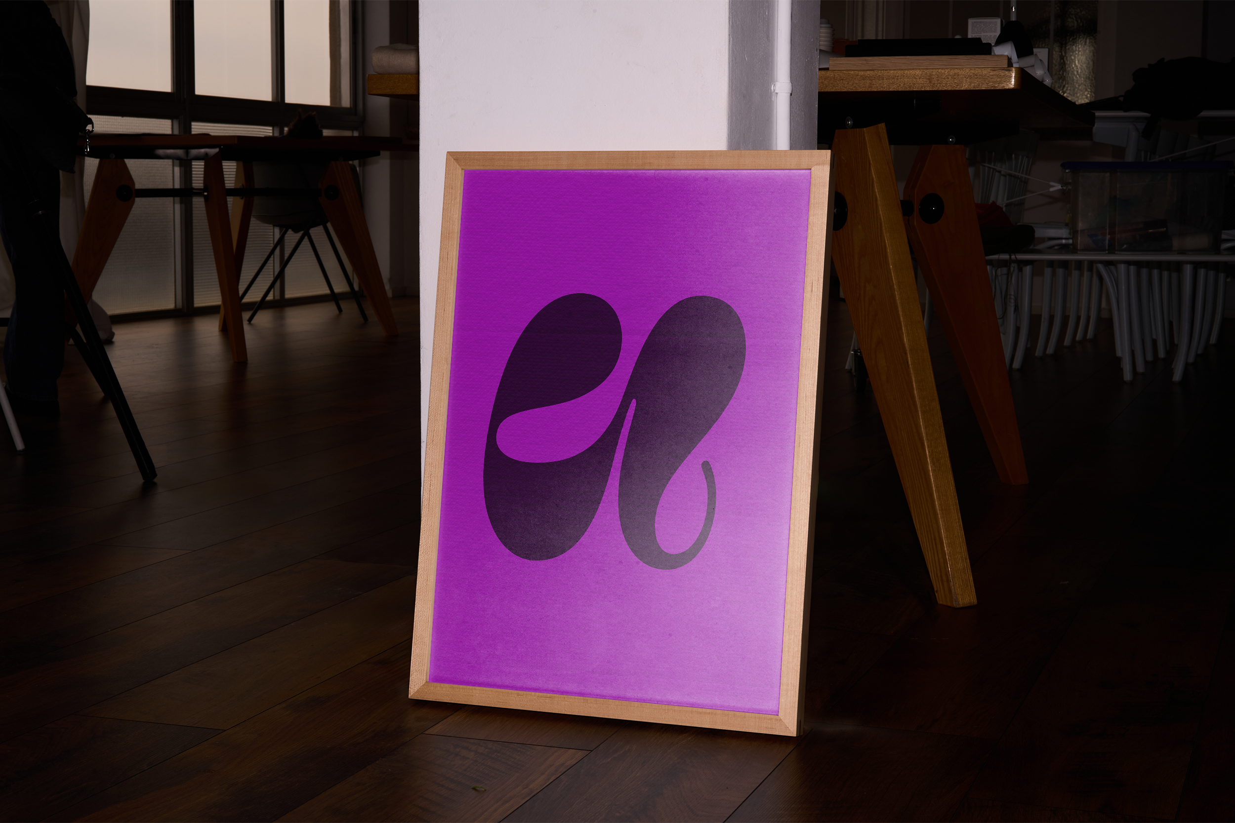 A light wooden framed poster with a large, swirling black graphic (resembling an abstract 'u') on a vibrant purple background. The poster is on a dark wooden floor in a dimly lit room.