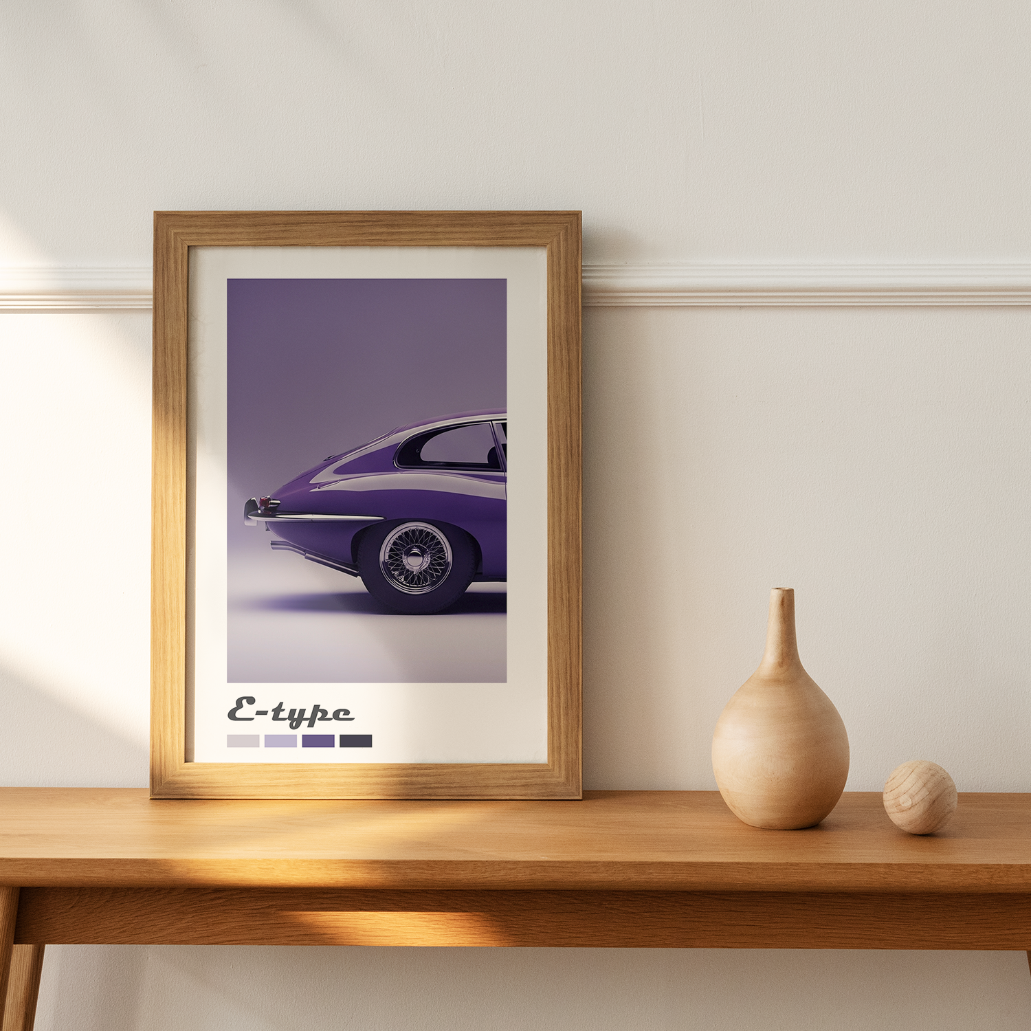 A wood-framed print featuring a close-up rear view of a deep purple classic Jaguar E-type sports car with color swatches below, resting on a light wooden table next to a wooden vase.