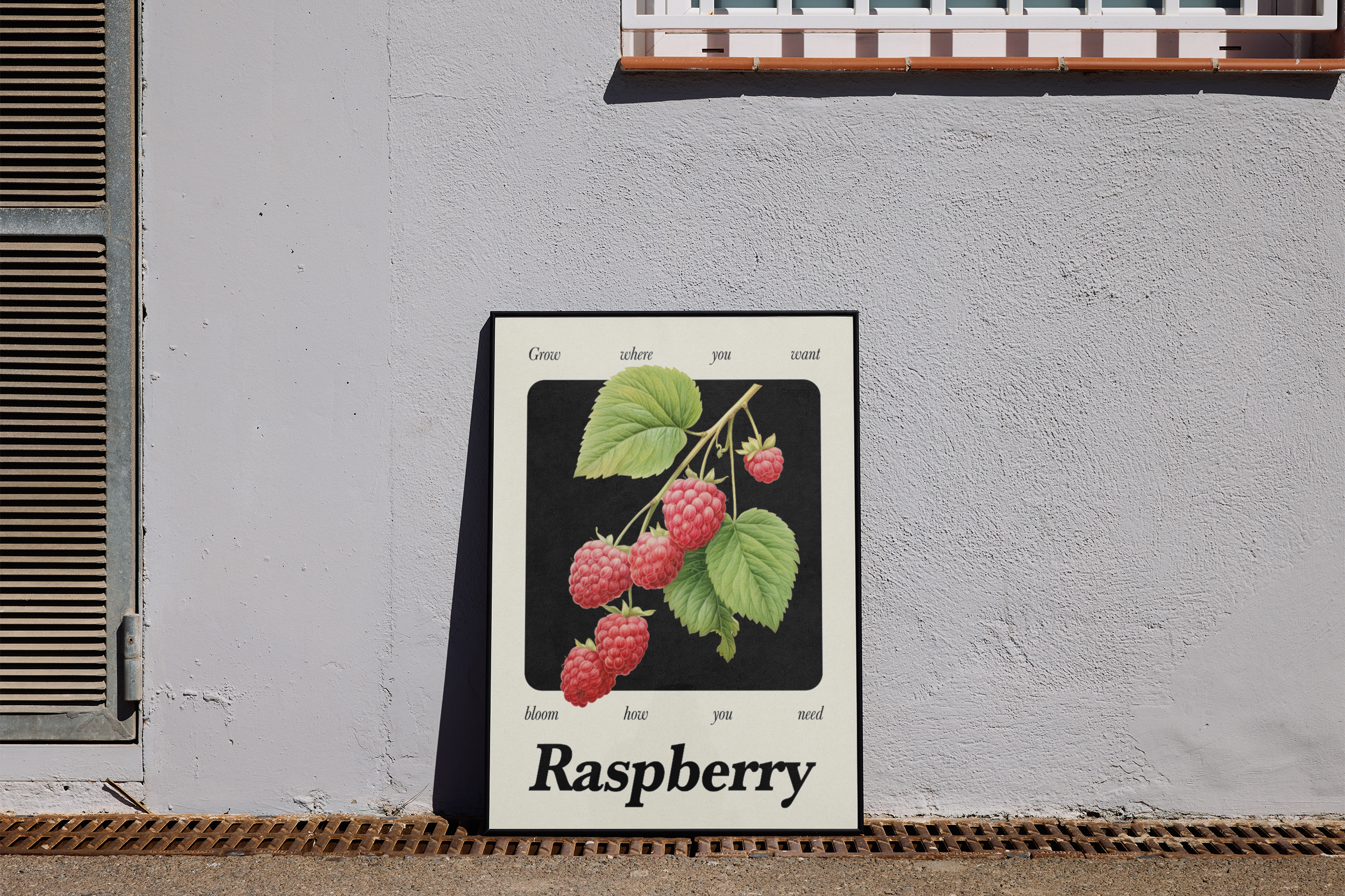 A black-framed print featuring a botanical illustration of raspberries and leaves on a branch against a black and white background with an inspirational quote, leaning on the ground against a light gray concrete wall.