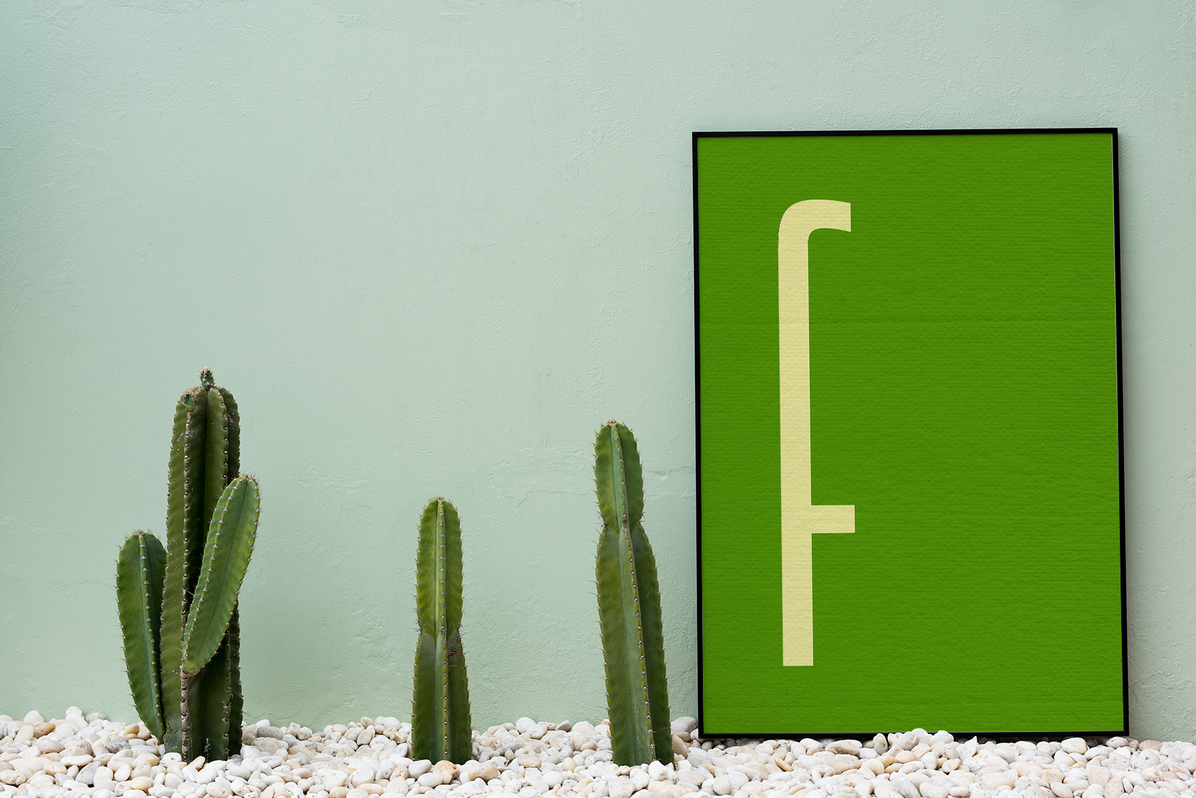 A framed art print displaying a thin, beige lowercase letter 'f' on a deep green background, propped up next to tall cactus plants and white pebbles.