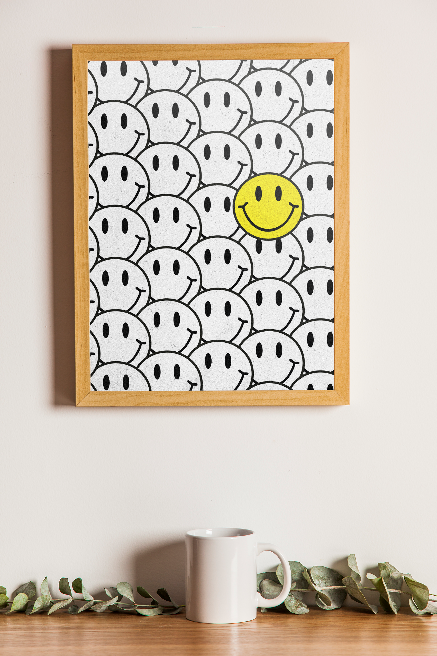 A wood-framed poster with a busy, crowded pattern of black and white outlined smiley faces, highlighted by one contrasting yellow smiley face, hanging above a small white mug.