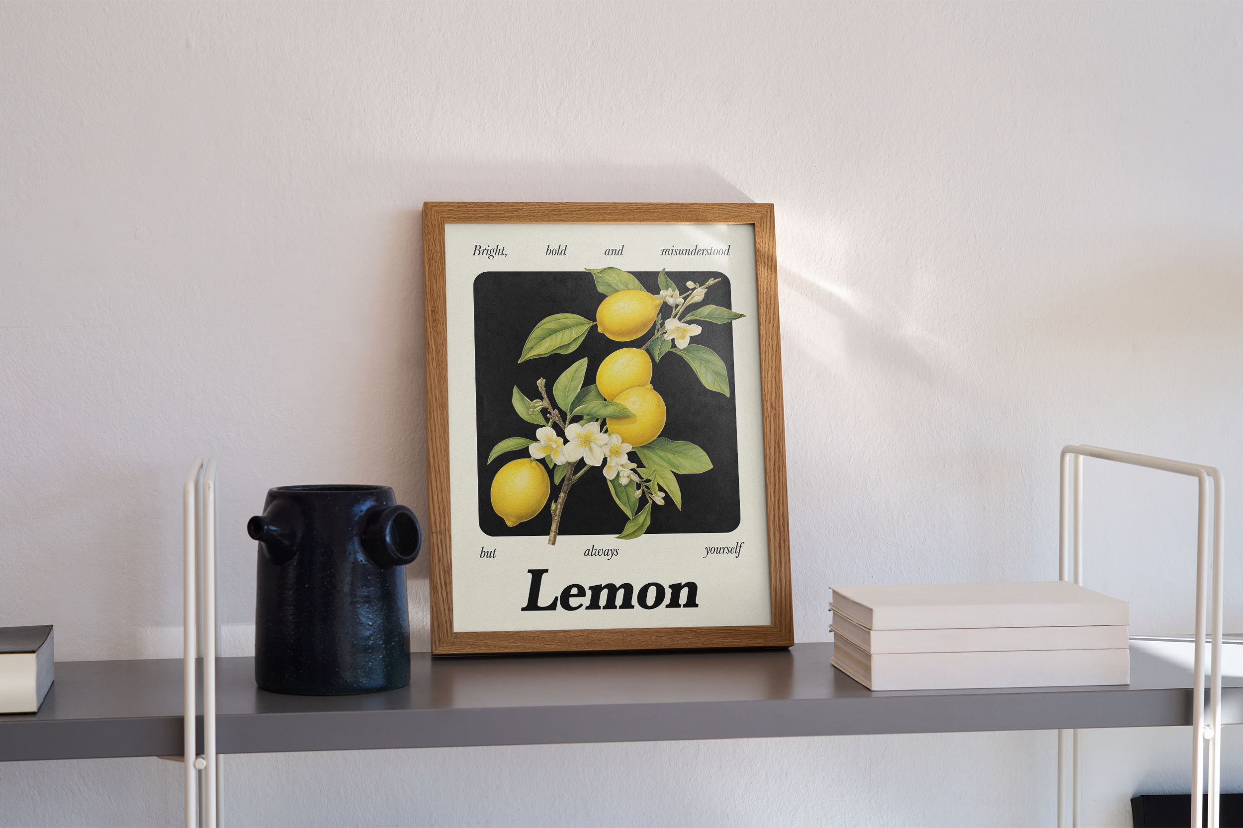 A wood-framed print on a grey shelf with an illustration of lemons on a branch, next to a black ceramic jar and a stack of white books. The print has the text "Lemon" at the bottom.