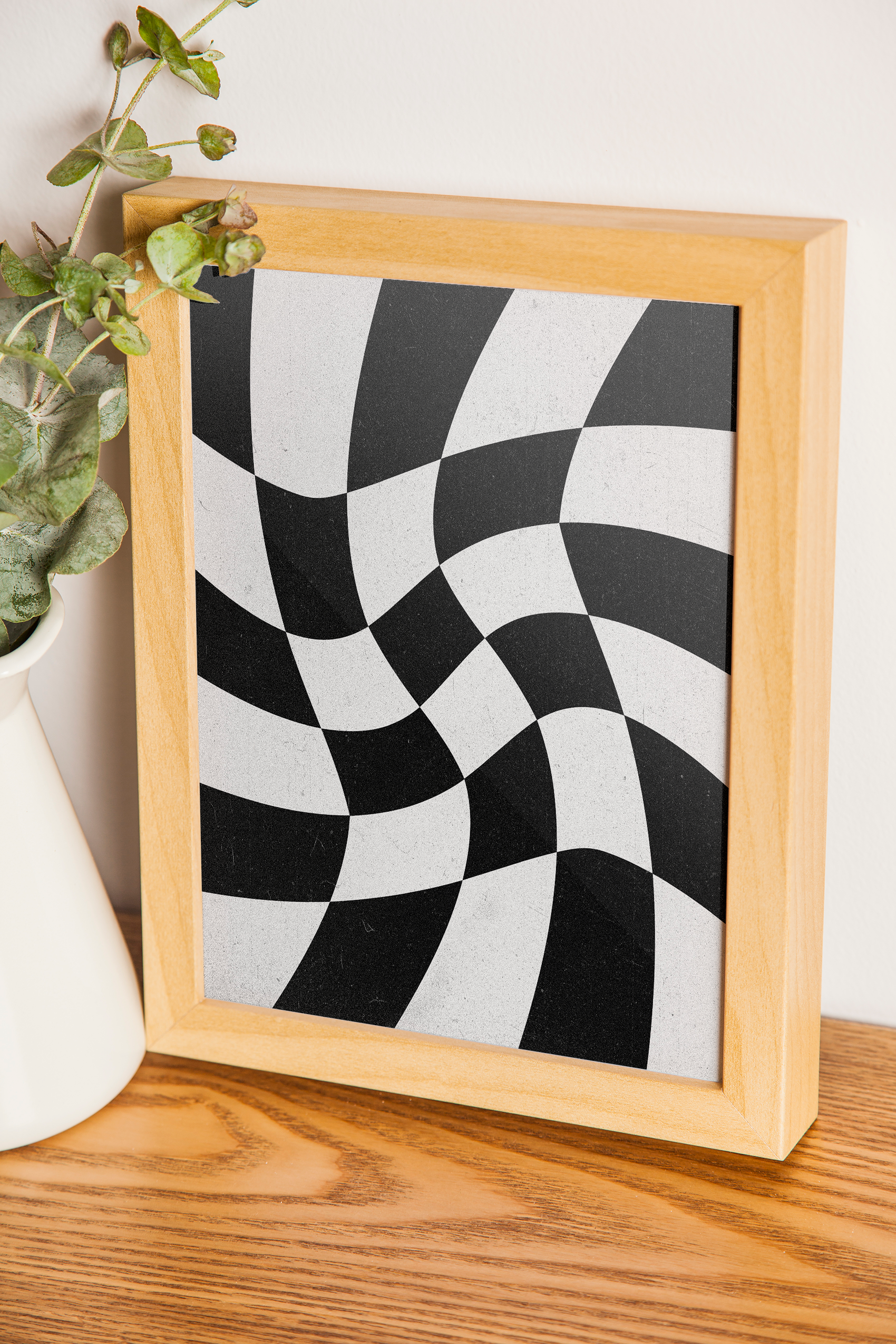 A vertical framed print with a wavy, black and white checkered pattern, resting on a wooden surface next to a white vase and green plant cuttings.